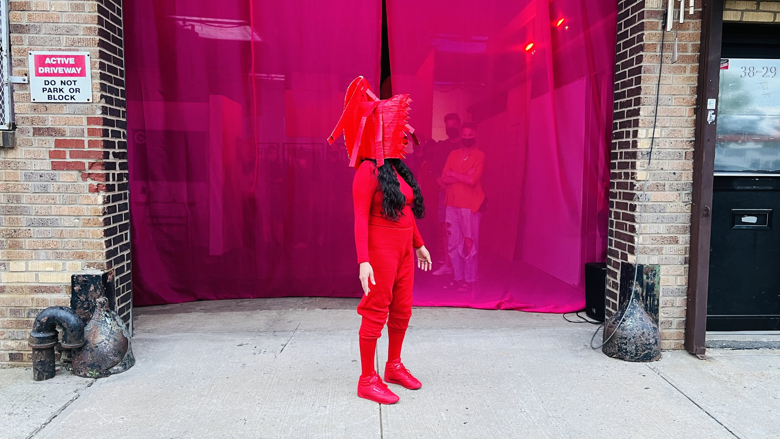 A performer dressed in red from head-to-toe and wearing a red mask with sharp points, stands in front of a billowing pink curtain outisde of a large rolling gate. Photo by Brian Rogers.