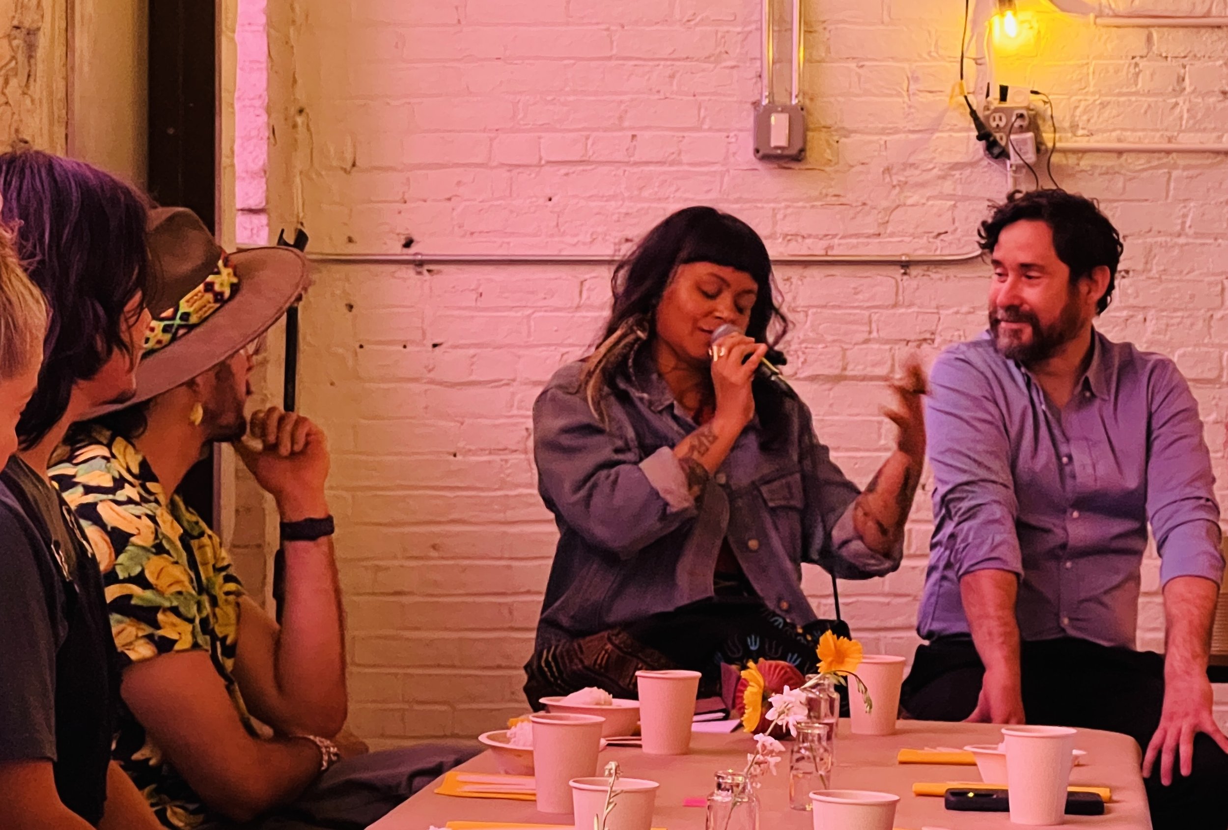 Two people at the end of a communal table are speaking. One is holding a microphone and gesticulates and the other listens. An audience is seated around them. Photo by Brian Rogers.