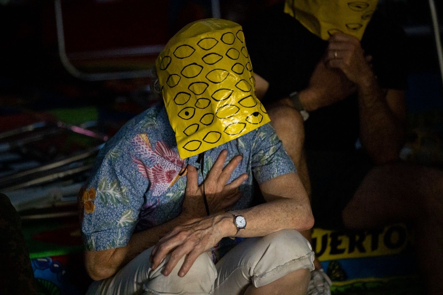 An audience member wearing a yellow papier-mache mask with multiple eyes drawn on it, places their palm on their chest. Photo by Maria Baranova.
