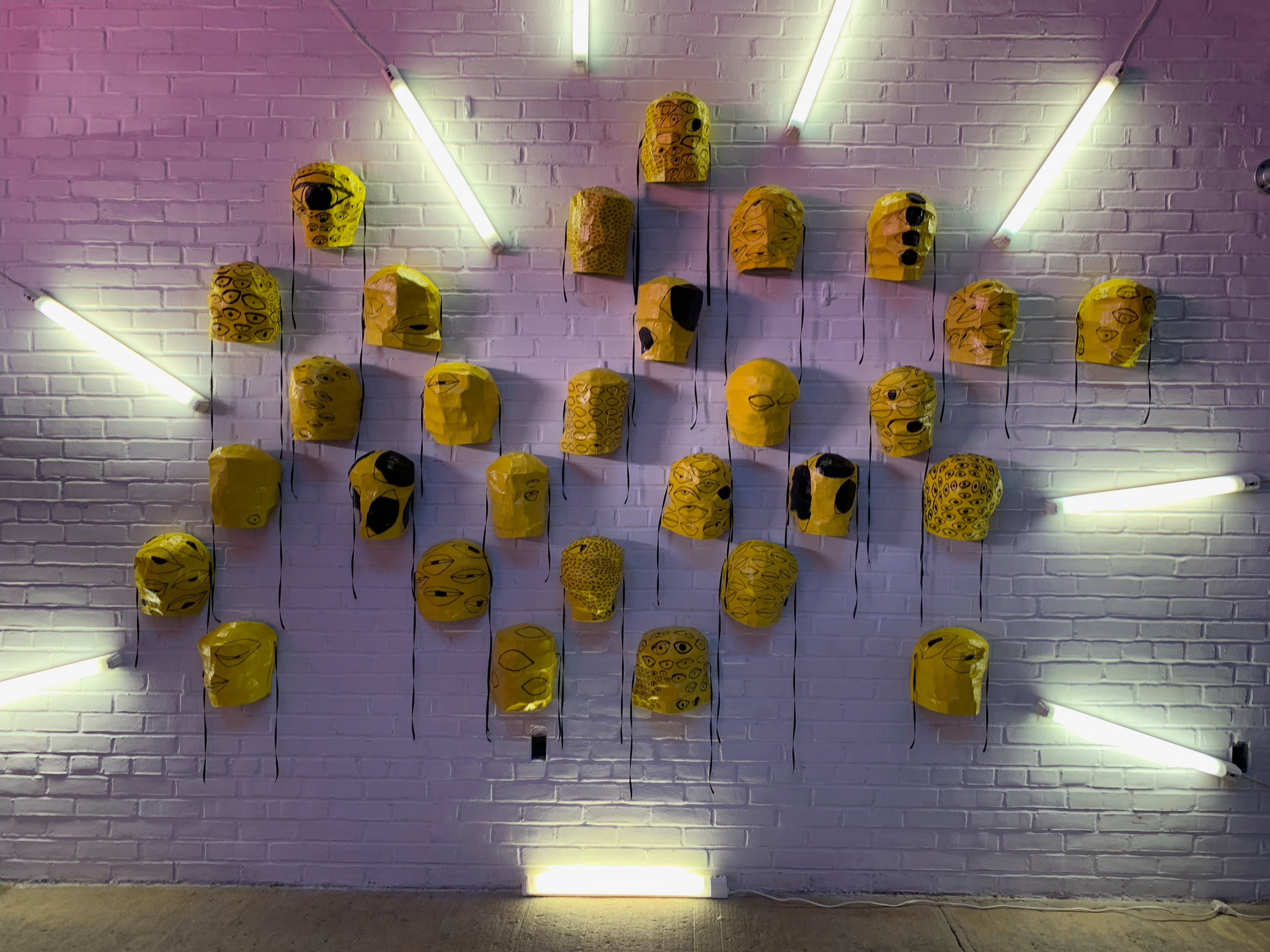 Thirty-two yellow papier-mache masks with black eyes drawn on them are hung on a white brick wall. They are lit by fluorescent lights radiating around them. Photo by Yanira Castro.