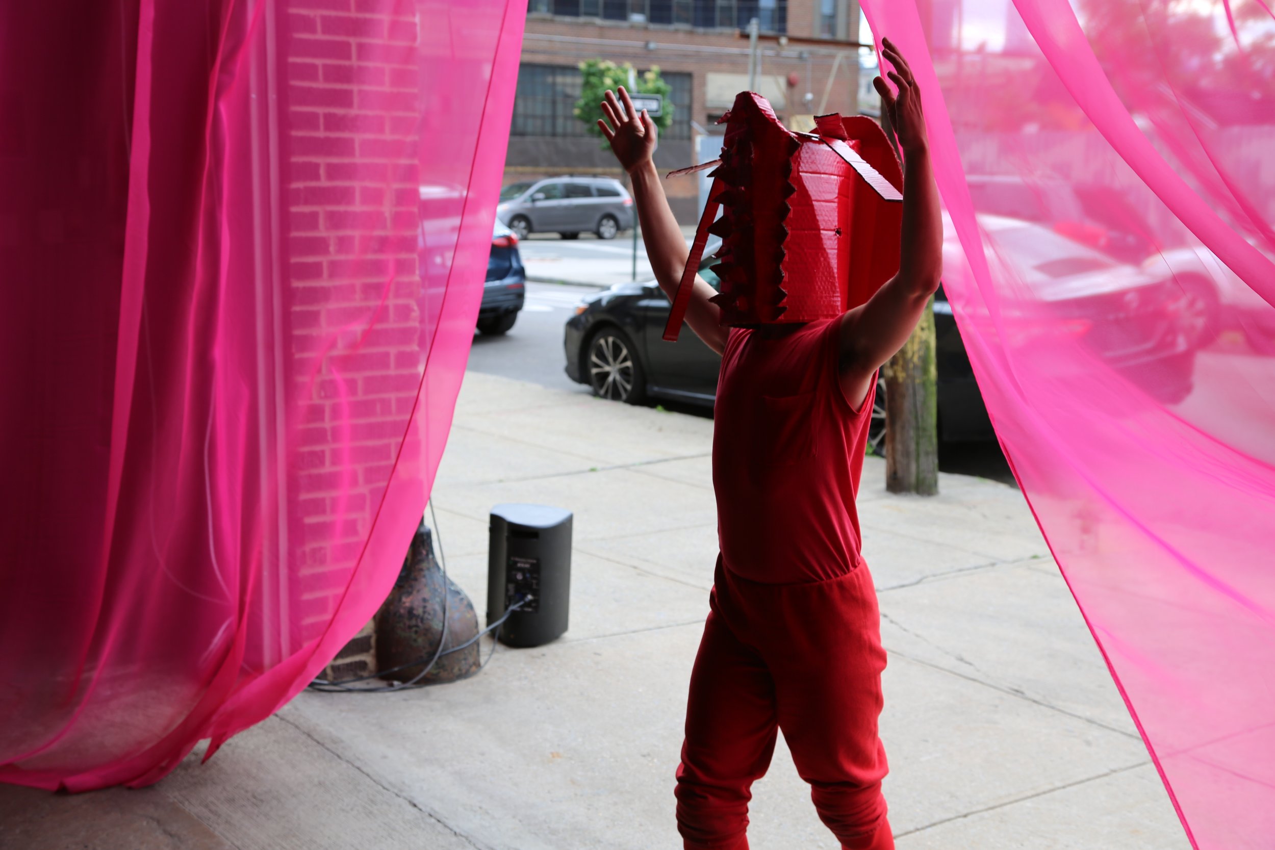 A performer dressed in red and wearing a large, red mask with sharp points covering the front with long tentacles at top, raises their arms and moves through a pink curtain. Photo by Brian Rogers.