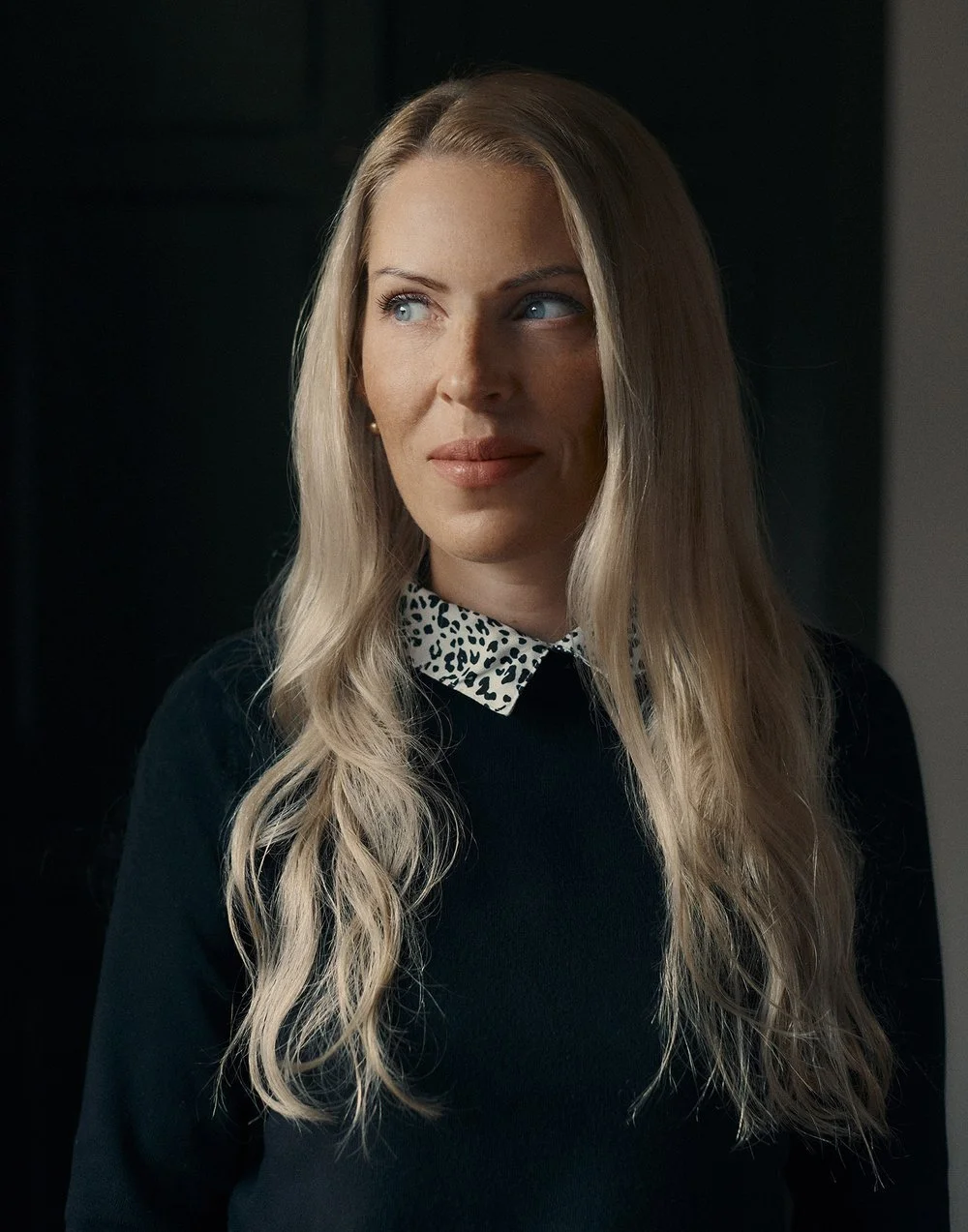 A woman with long blonde hair and blue eyes, wearing a dark top with a leopard print collar, looking to the side.