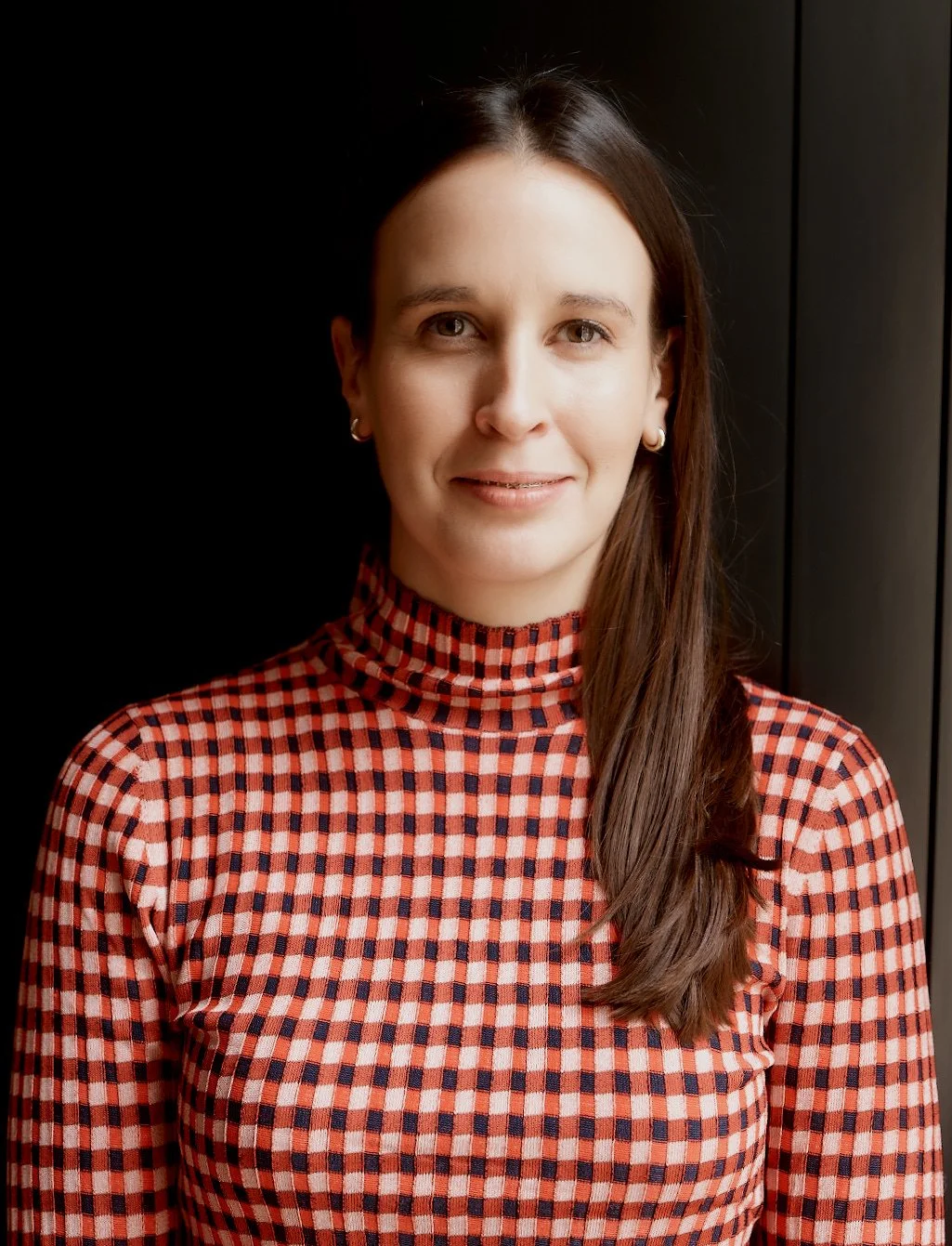 A woman with long brown hair wearing a checkered red and black turtleneck, standing indoors next to a black wall.