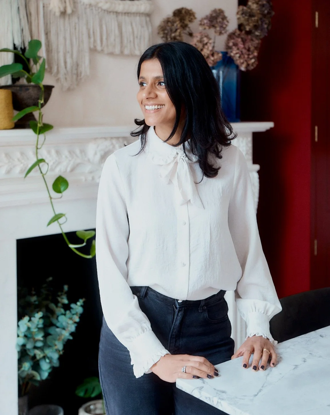 Woman with dark hair smiling, standing in a room with plants and a white fireplace.