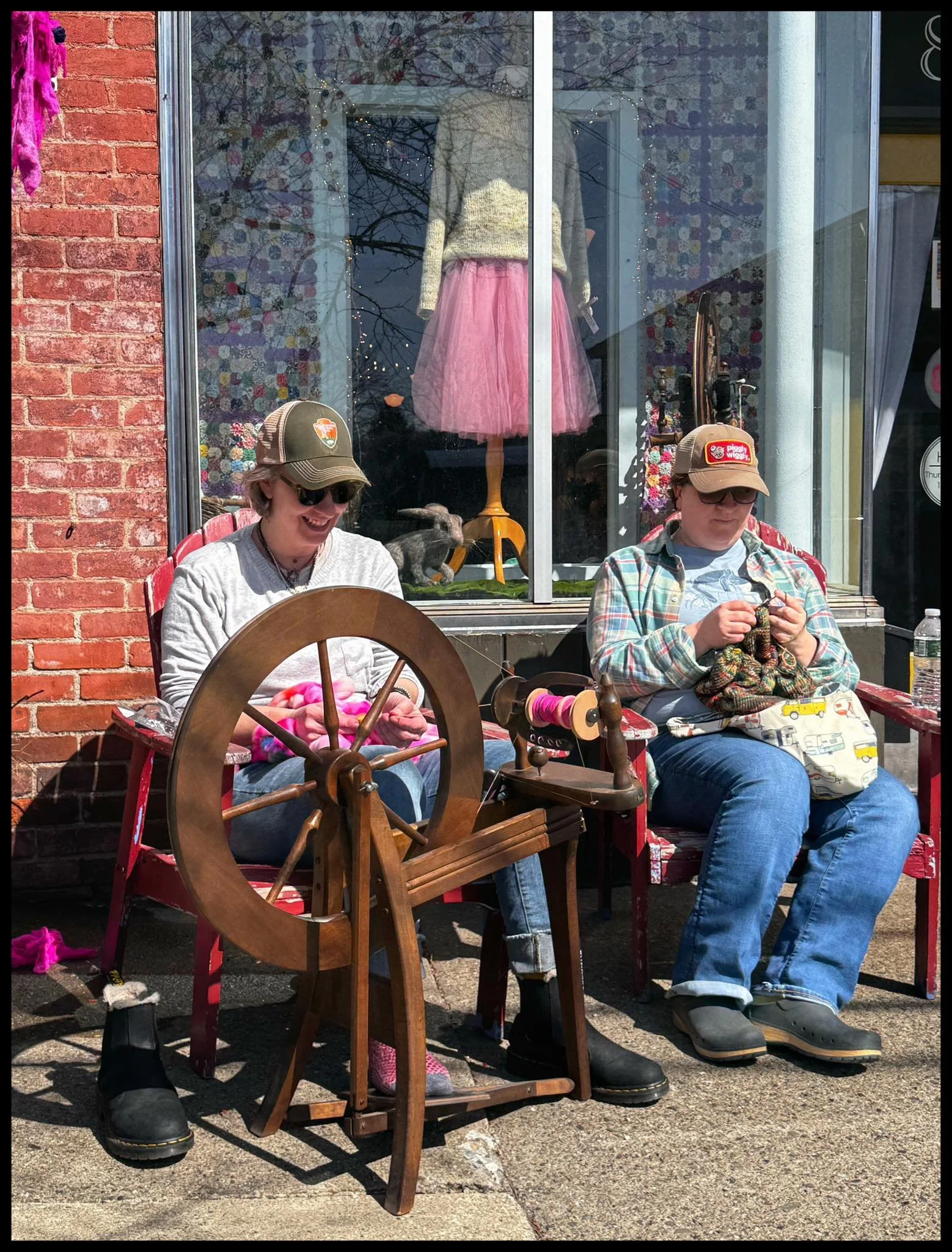 Two women wearing baseball caps and smiling are sitting outside on a sunny day. One is spinning yarn on a brown wooden spinning wheel and the other is knitting. The background is a brick storefront with a large window.