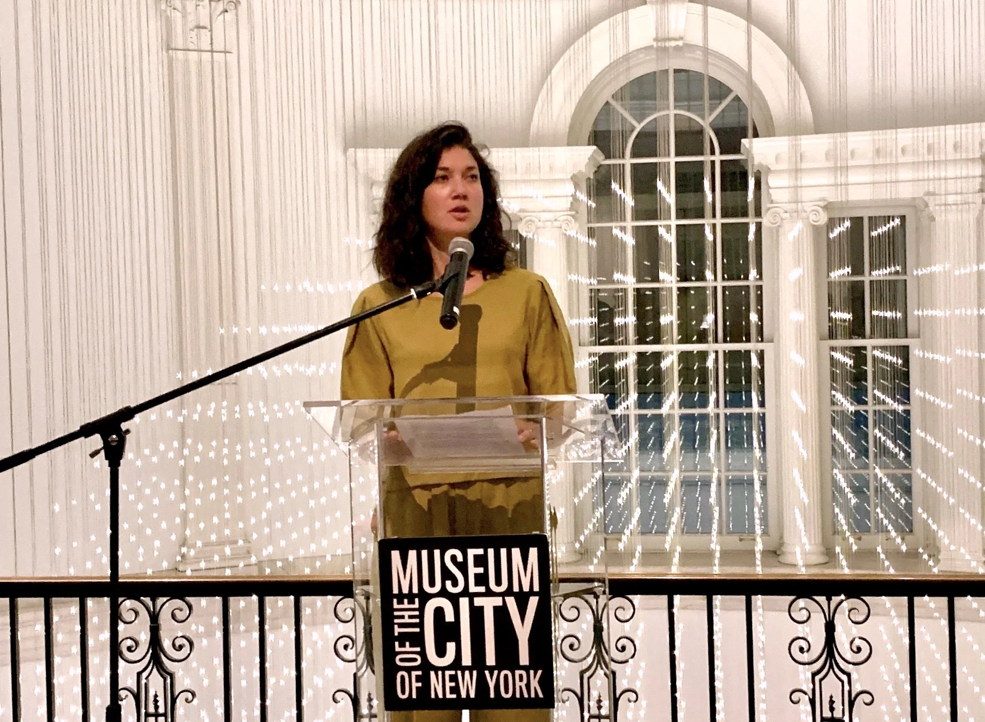 A woman with dark curly hair standing at a clear podium with a microphone, speaking at the Museum of the City of New York. Behind her is a large arched window and decorative white walls.