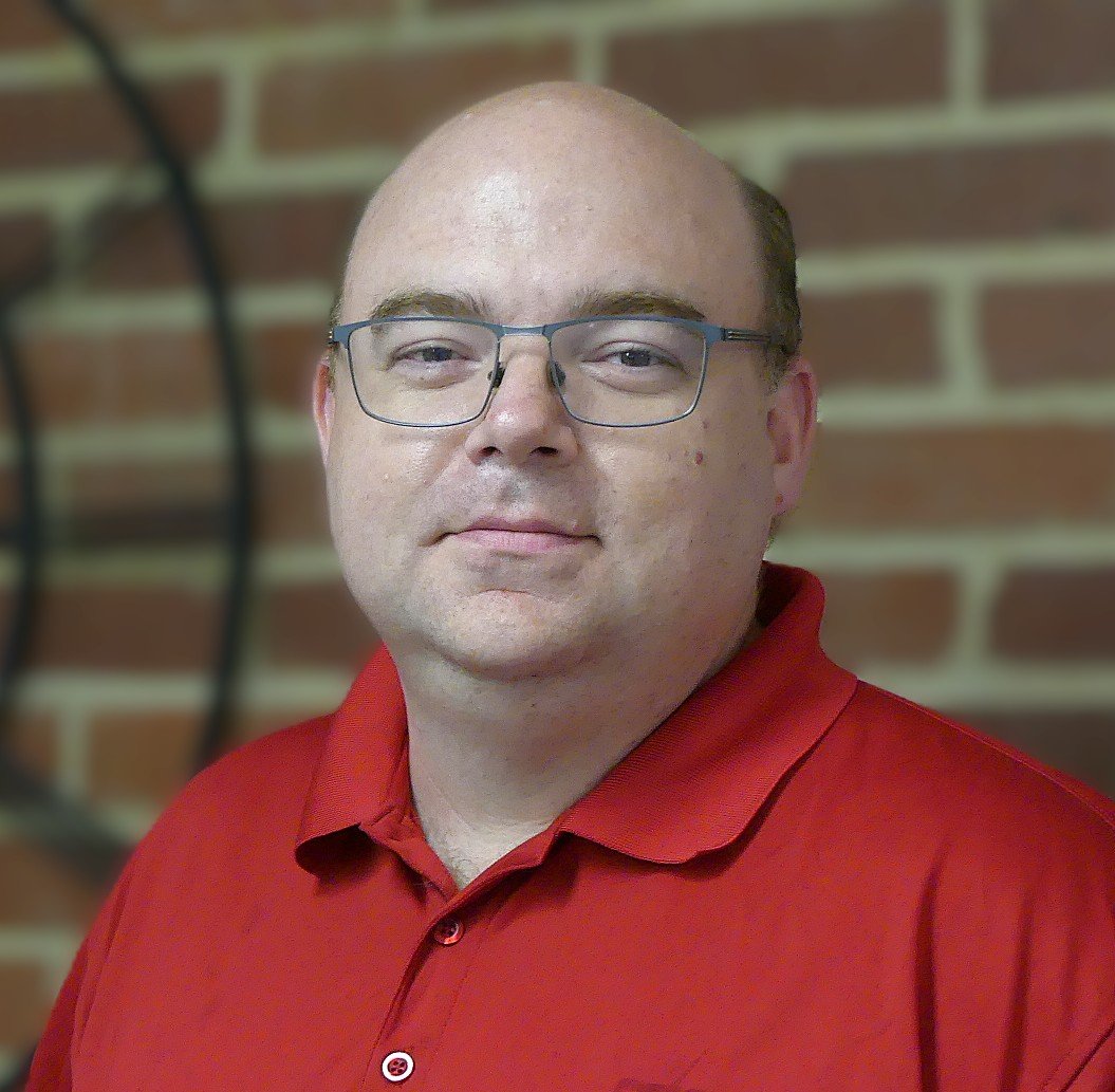 Smiling man in glasses wearing a light blue collared shirt, standing in front of a brick wall.