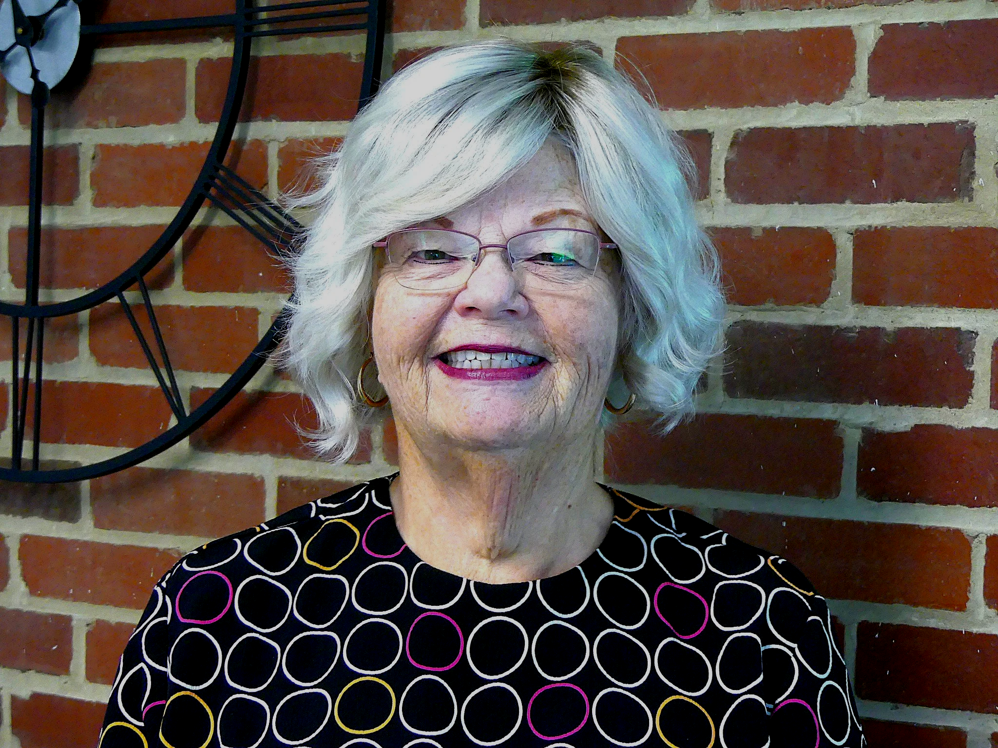 Smiling person with braided hair, wearing a colorful floral top and white cardigan, against a brick wall background.
