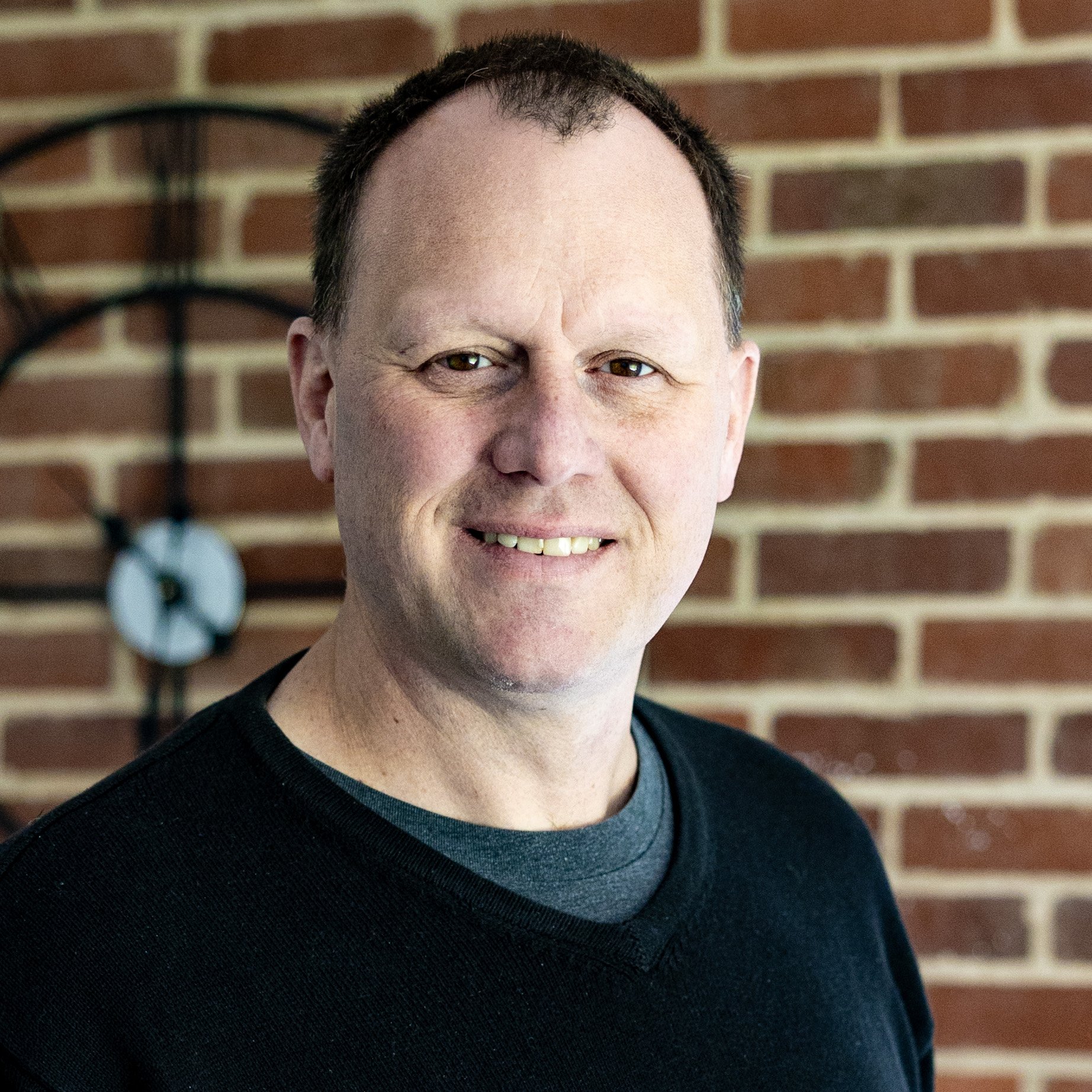 Man with glasses and a beard smiling in front of a brick wall.