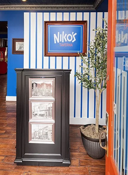 Photo of the entrance to a vibrant Greek restaurant with a blue and white pinstriped wall, host stand, and olive tree behind a bright orange door.