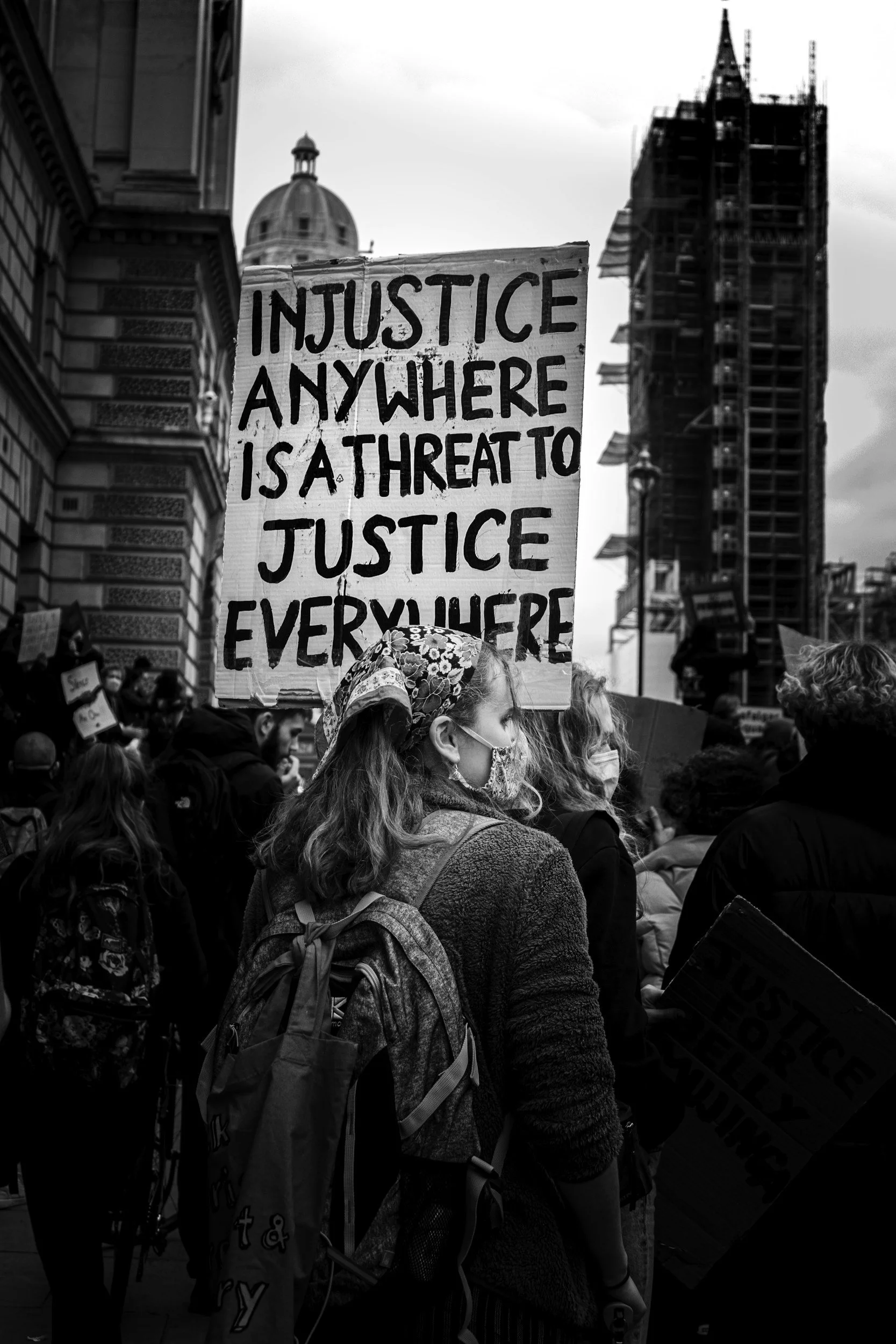 A crowd of people gathered at a protest or demonstration on a city street. A woman with a backpack and face mask is holding a sign that reads, 'Injustice anywhere is a threat to justice everywhere.' Buildings and a tower are visible in the background.