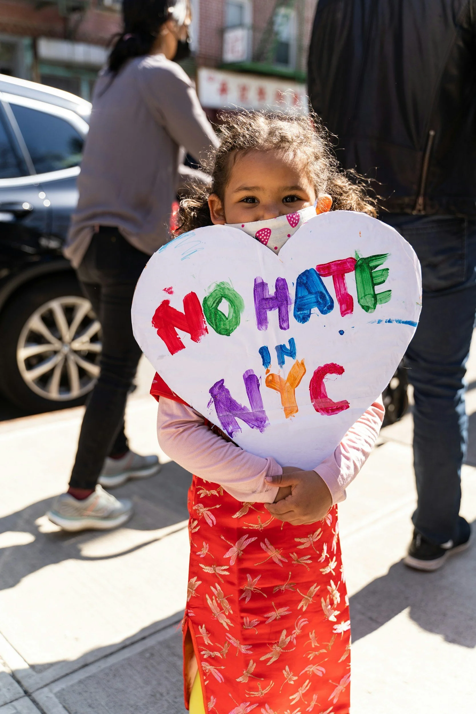 A young girl with curly hair, wearing a floral mask, holds a colorful heart-shaped sign that says 'NO HATE' in rainbow colors. She is outdoors, with sunlight on her face, and other people and cars are visible in the background.