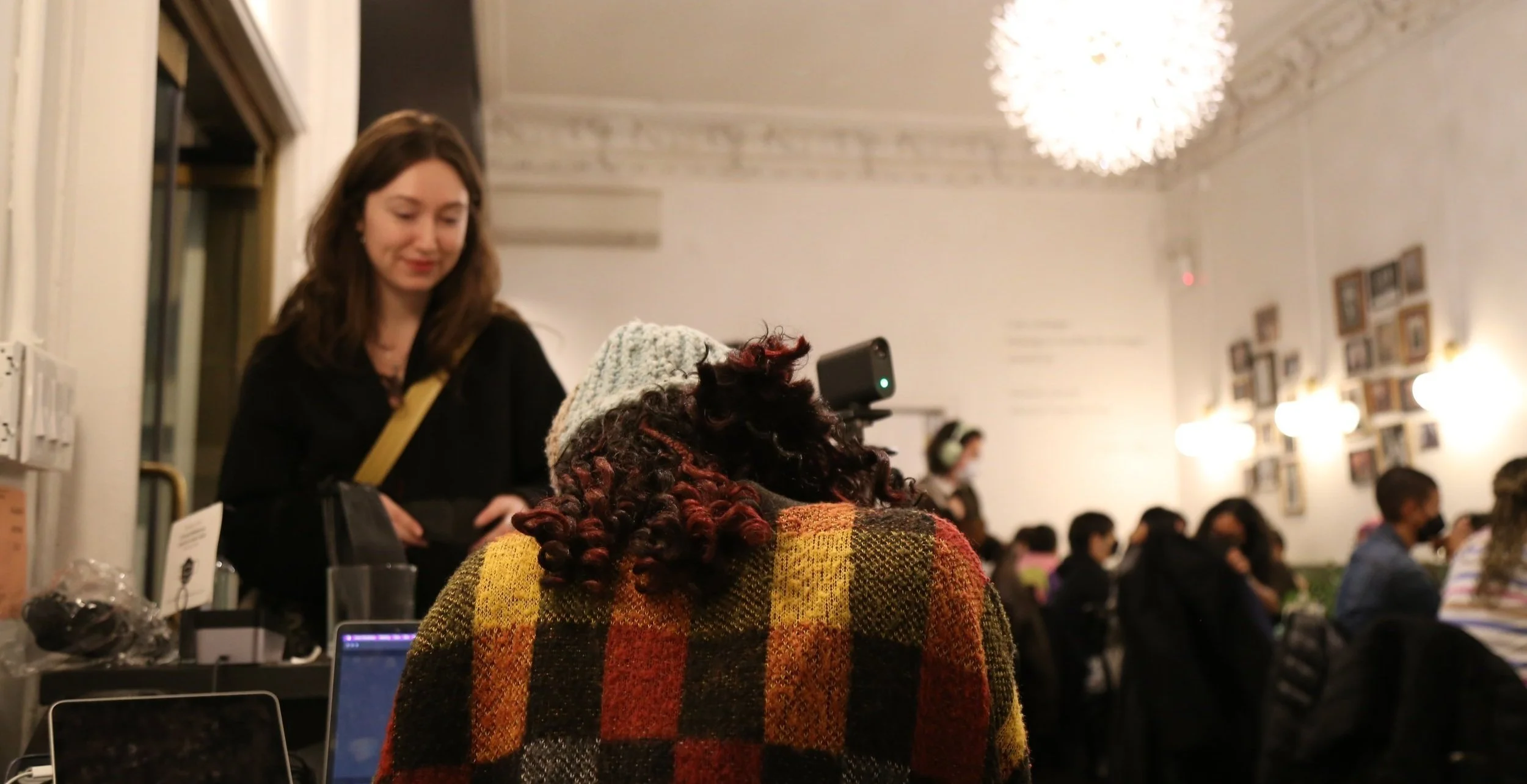 Group of people inside a cozy cafe with warm lighting, one woman in focus facing another person wearing a plaid sweater and a knitted hat.