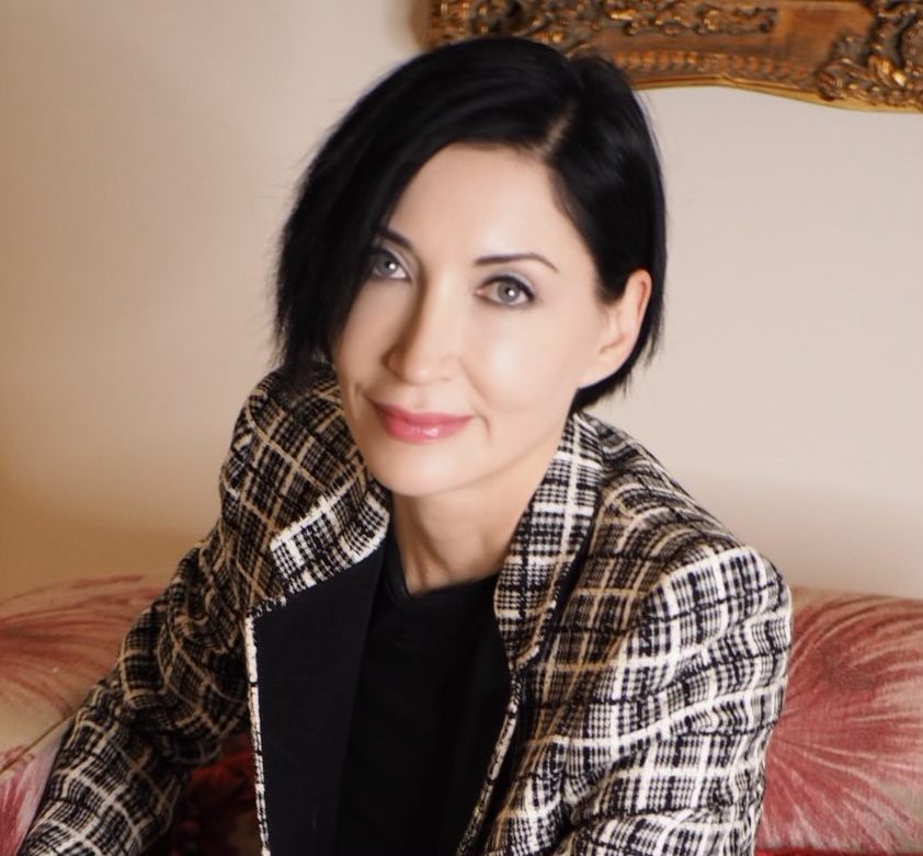 A woman with short black hair and light skin, wearing a checkered blazer, sitting indoors on a pink cushion with a gold decorative frame in the background.