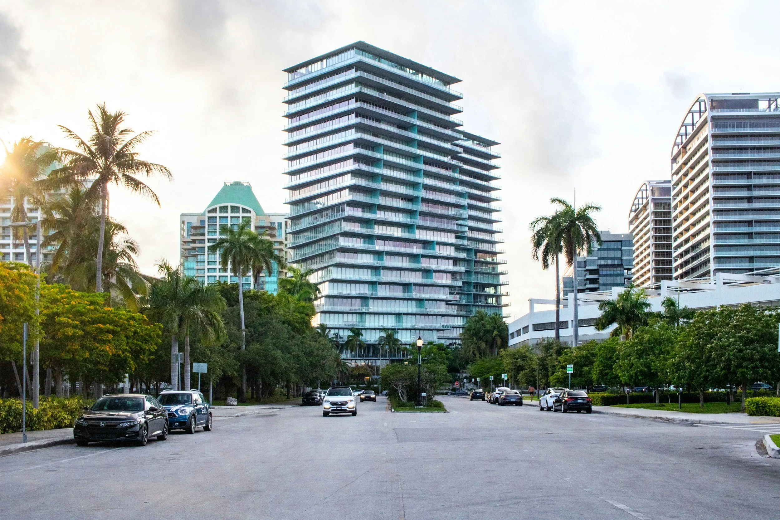 A cityscape with modern high-rise buildings, palm trees, and a parking lot with several cars during daytime.