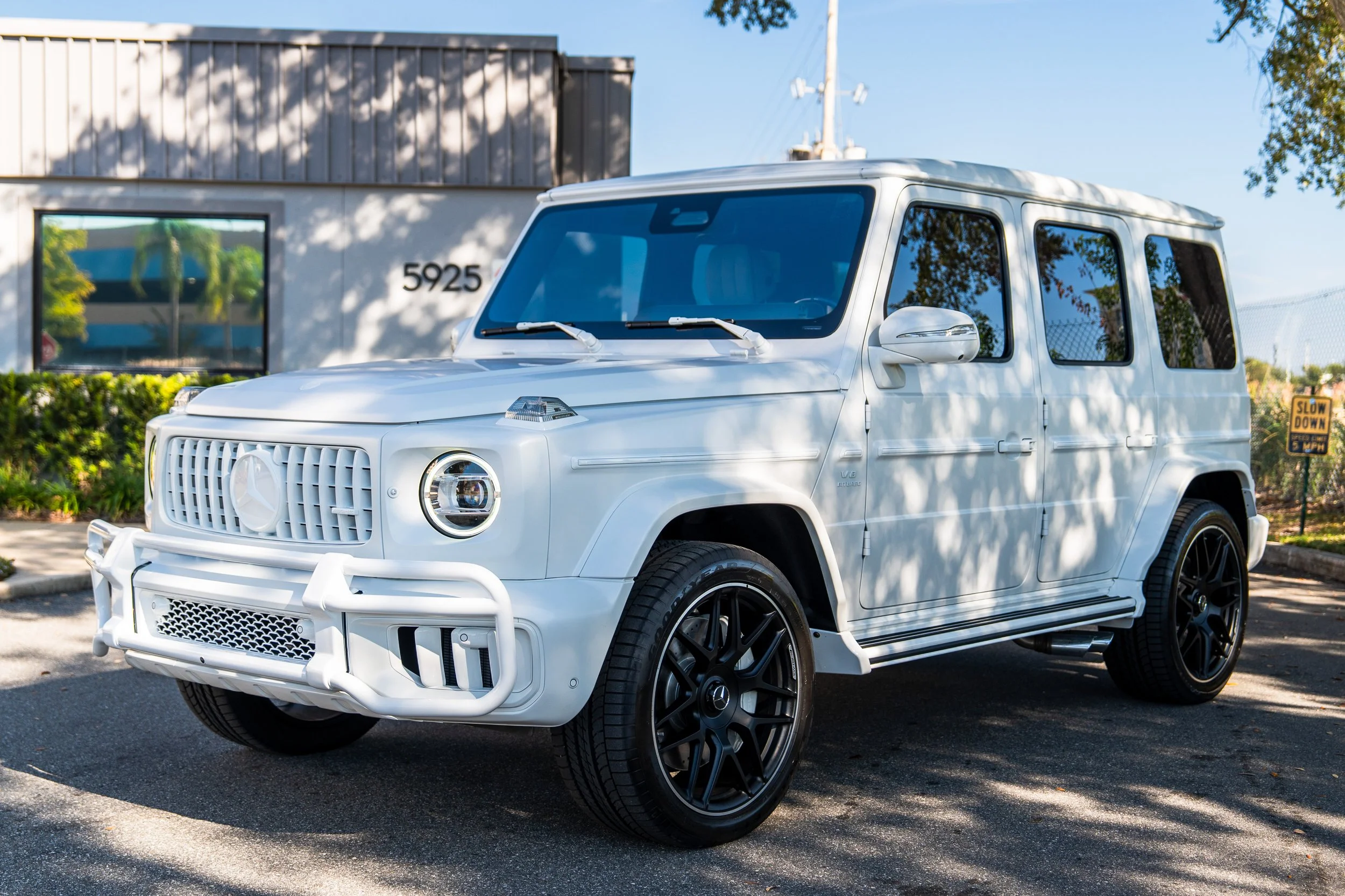 A white Mercedes-Benz G-Class SUV parked outdoors with trees and a building in the background.
