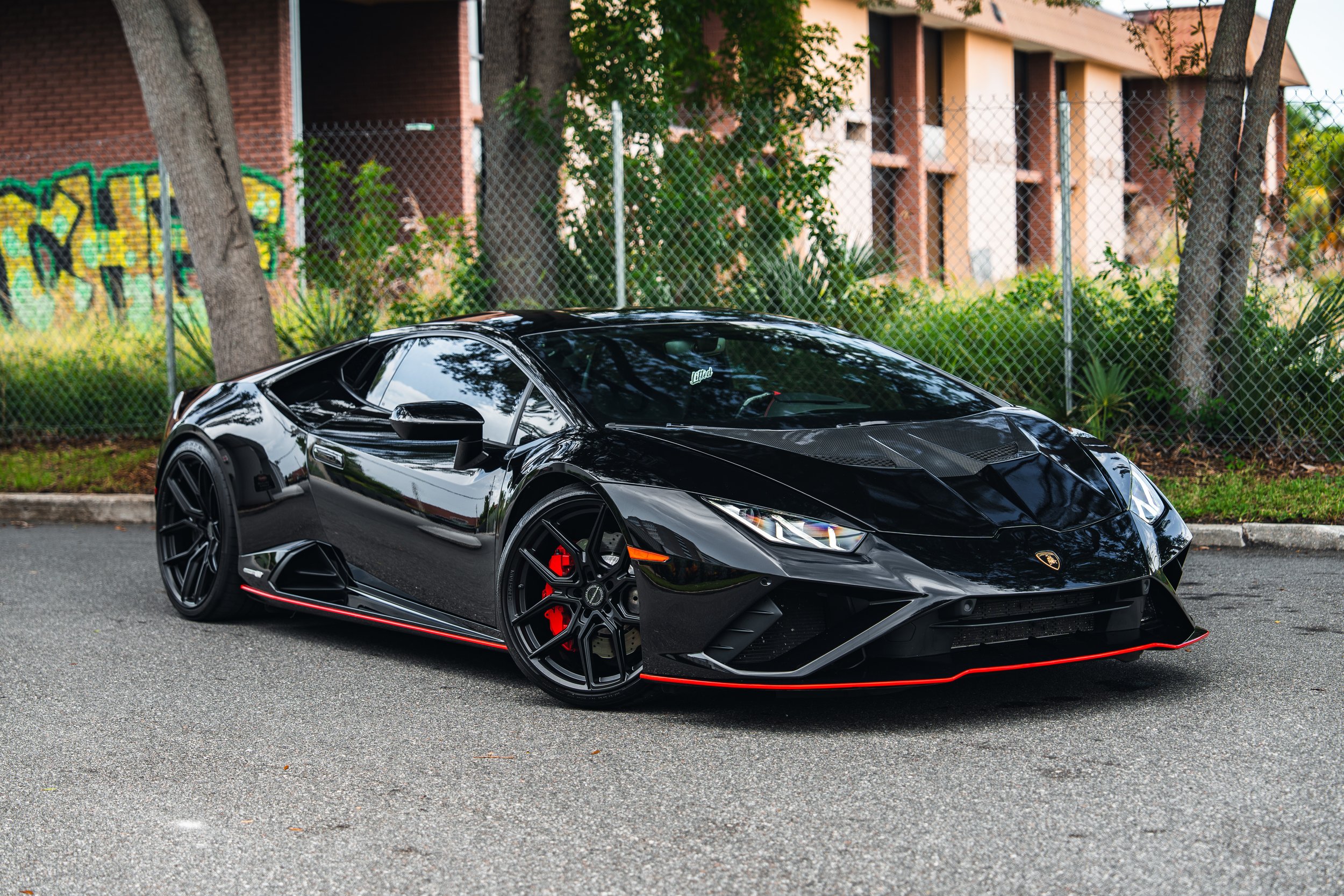 Black Lamborghini Huracan parked on street with trees and a chain-link fence in background.