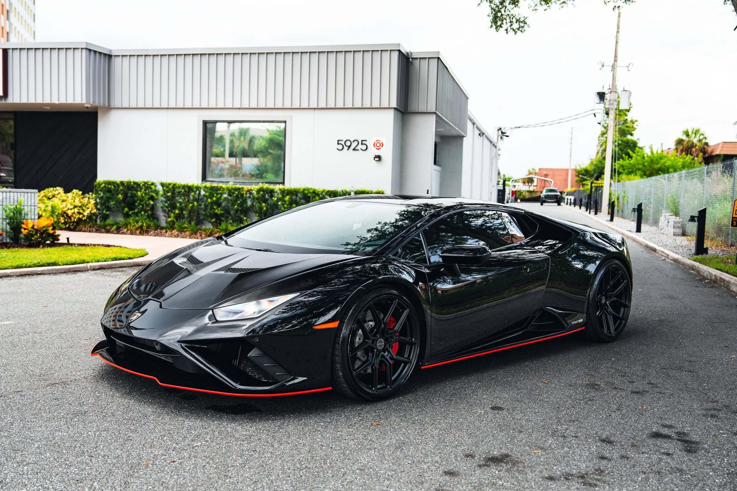 Black Lamborghini sports car parked in an urban area with modern buildings and green landscaping.
