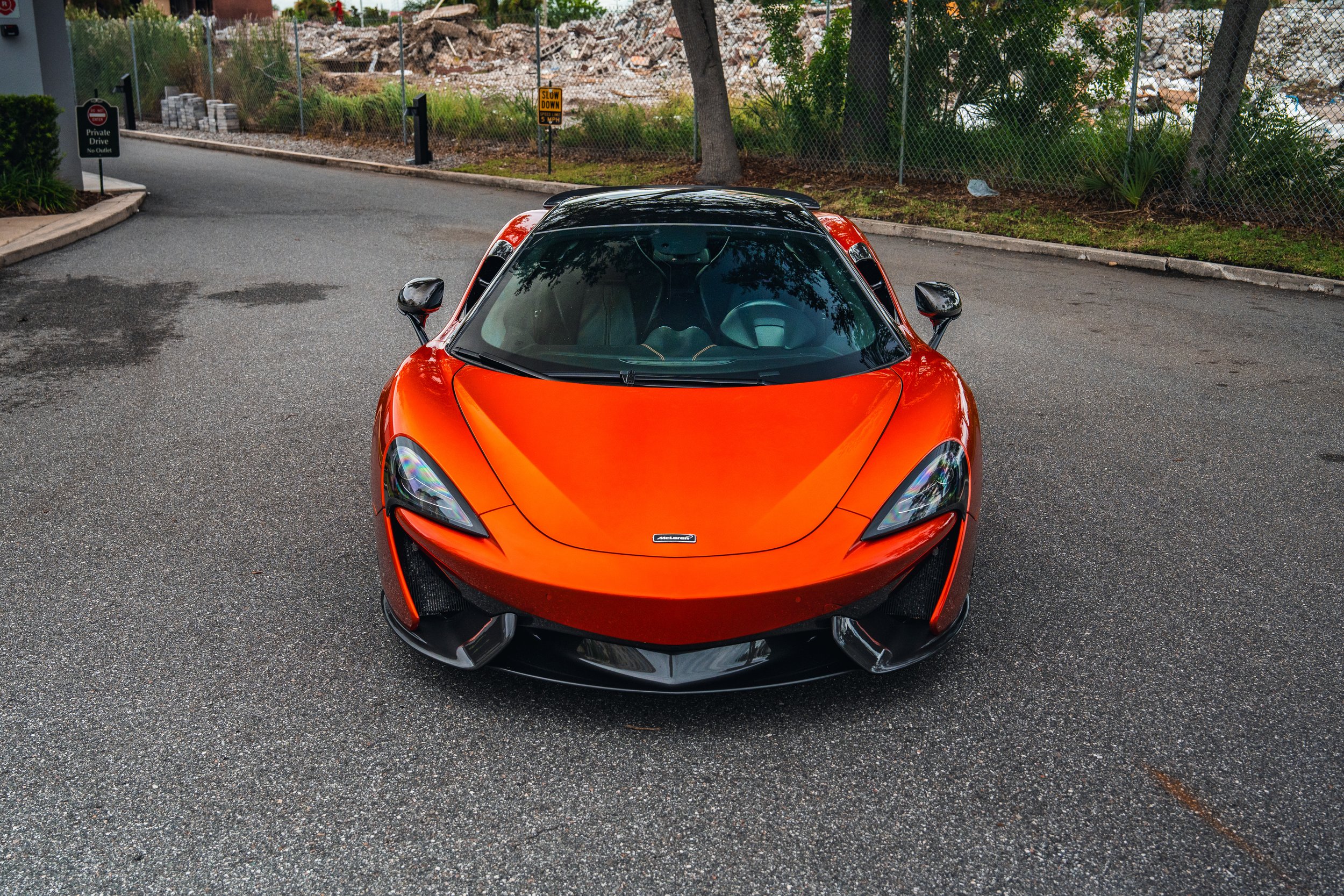 Front view of an orange sports car parked on a paved road with trees and a chain-link fence in the background.