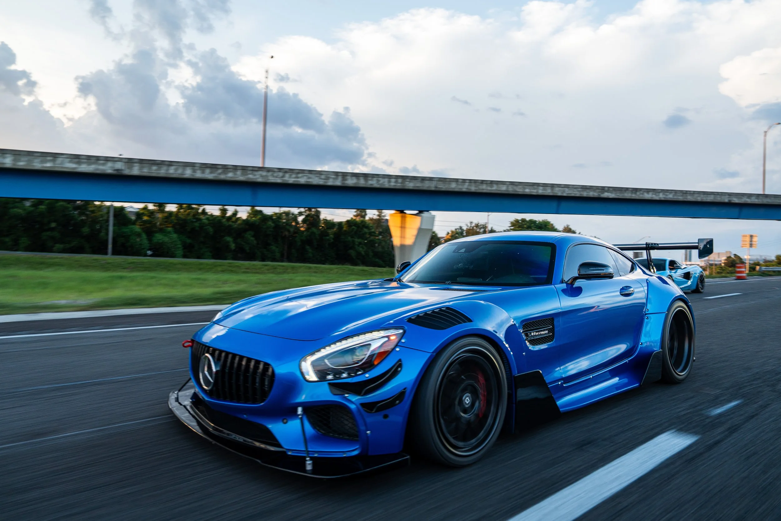 A blue Mercedes-Benz race car driving on a highway with a background of trees, a blue sky with clouds, and a second blue race car following behind.
