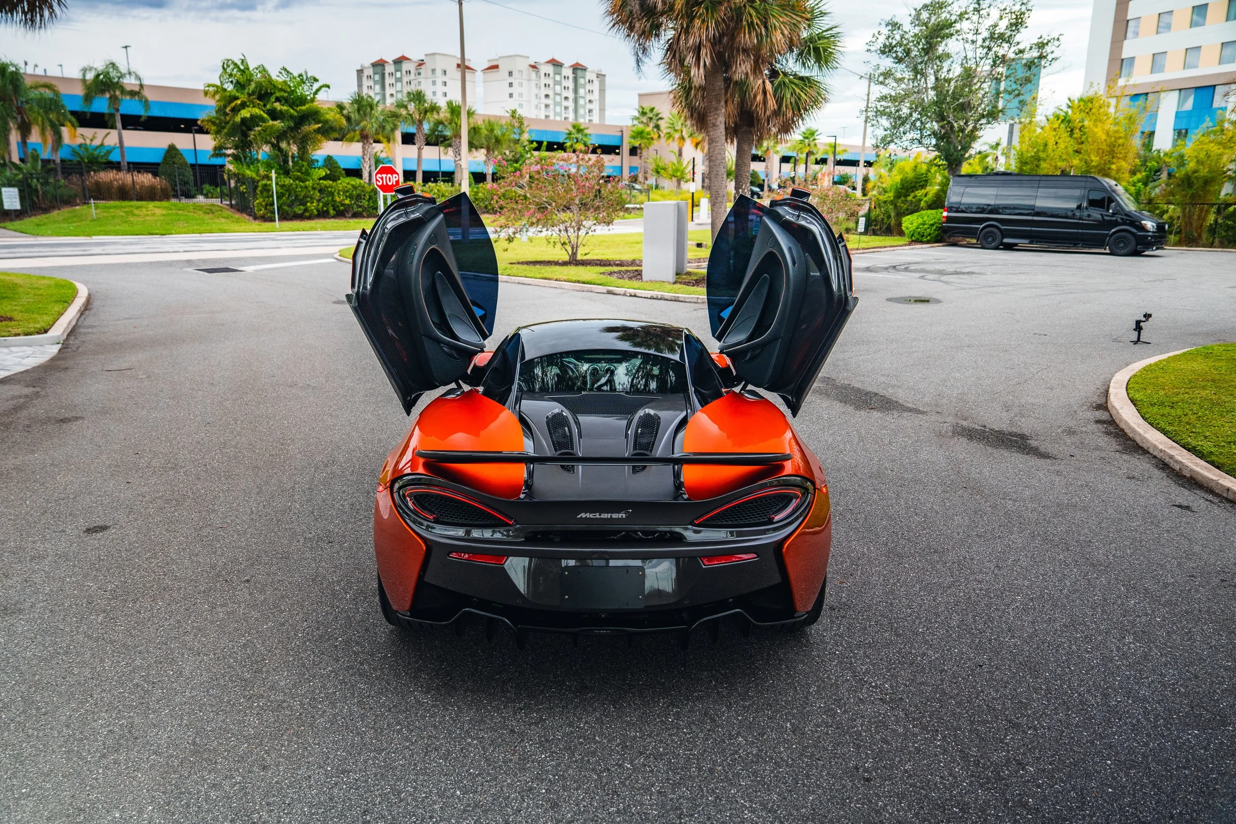 Red McLaren sports car with open gullwing doors parked on an asphalt driveway in a landscaped area with trees and bushes, background of multi-story buildings and a black van.