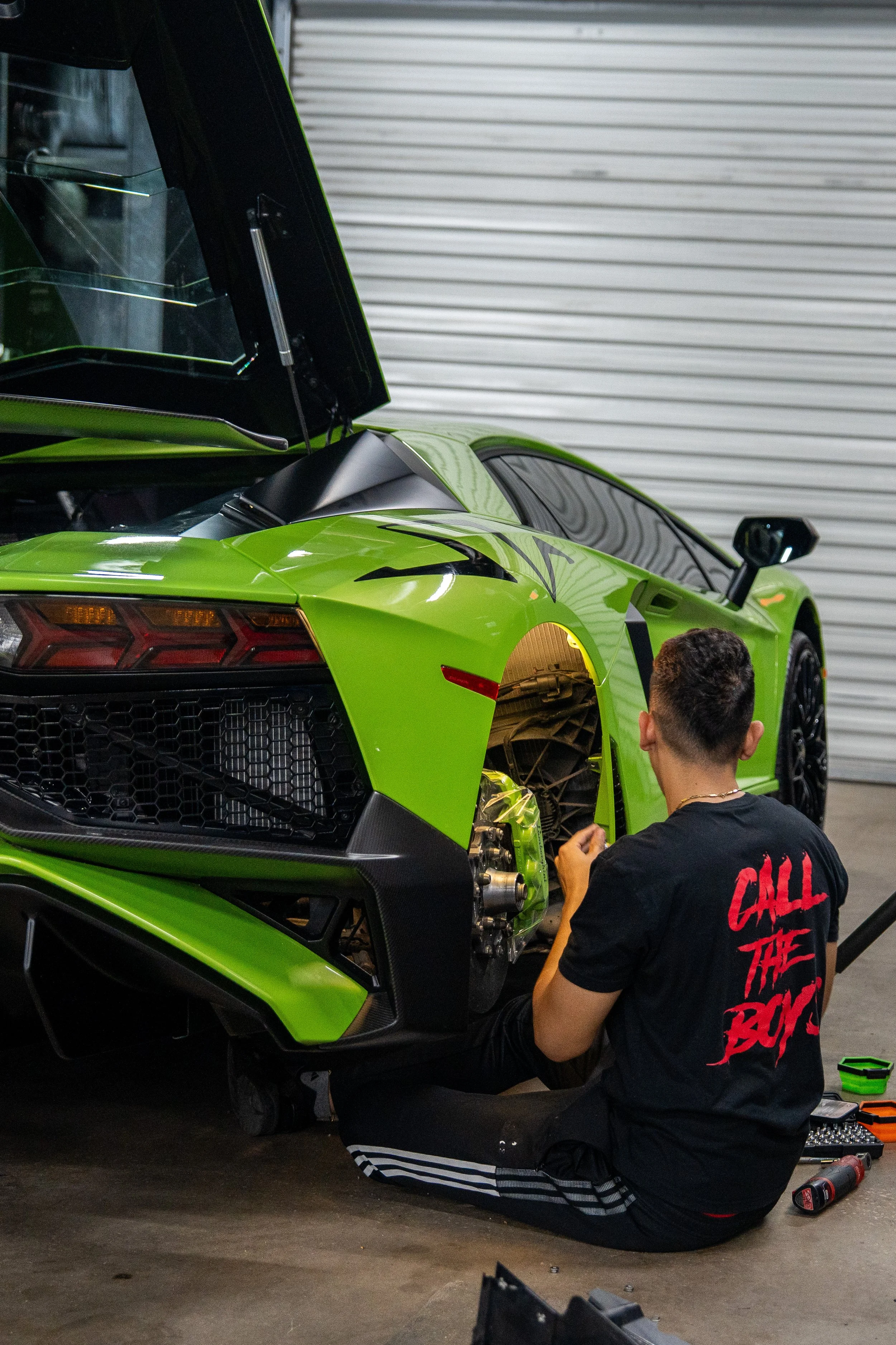 A person working on a lime green sports car inside a garage, with the rear wheel removed and the person sitting on the ground.