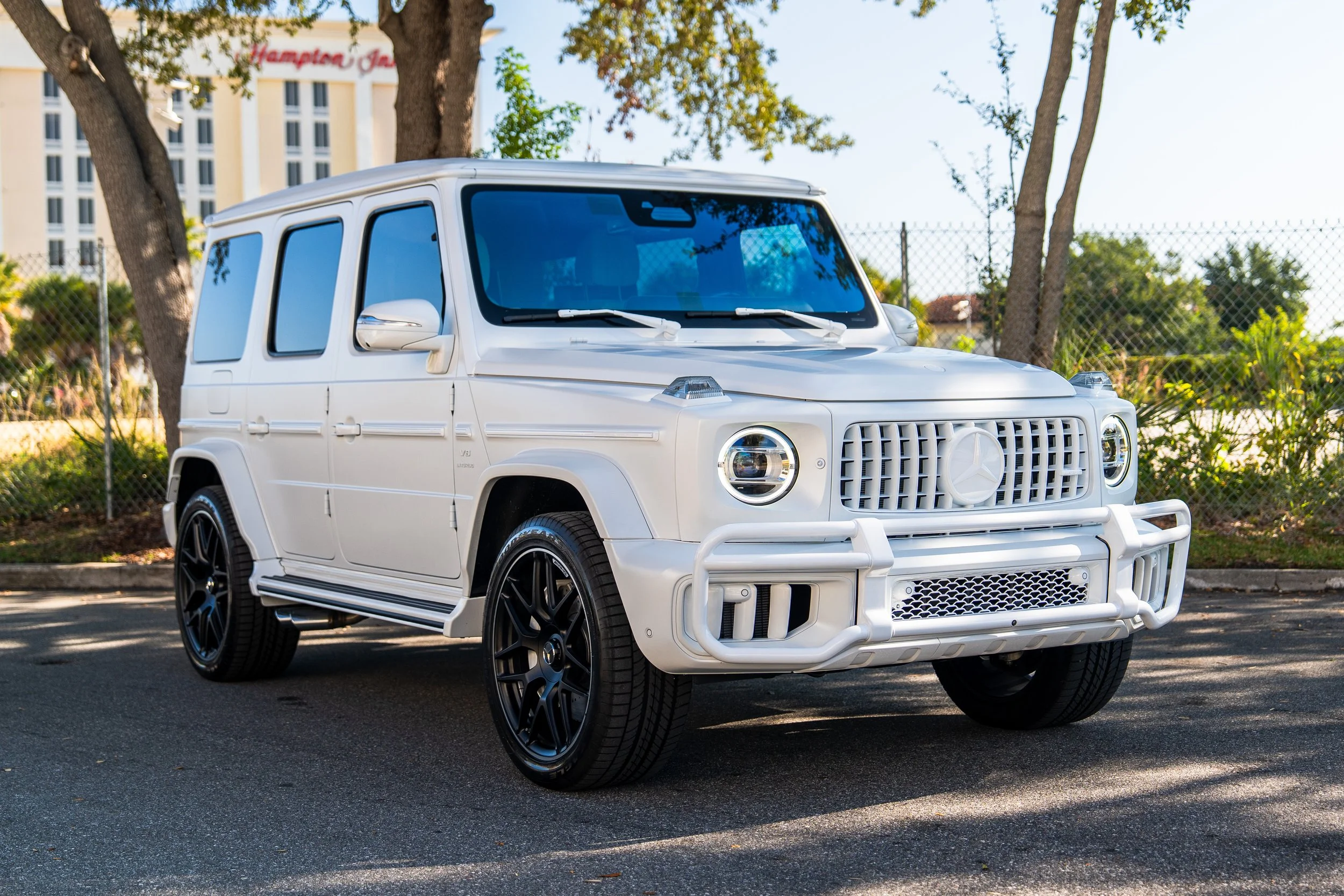A white Mercedes-Benz G-Class SUV parked on a street near trees and a chain-link fence, with a Hampton Inn hotel in the background.