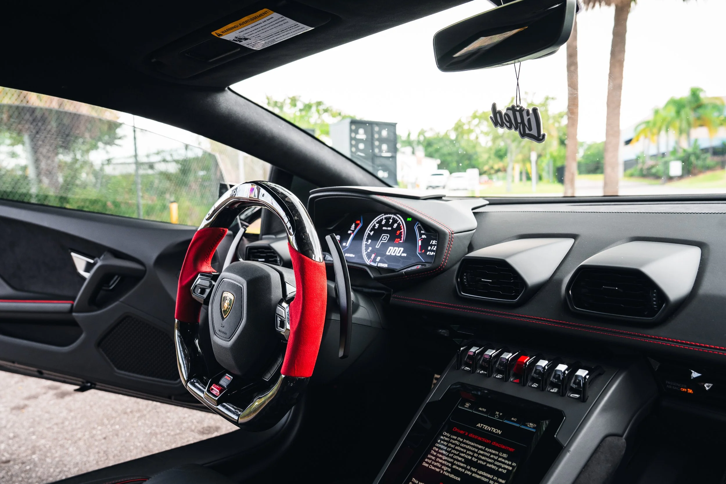 Interior of a luxury sports car, showing the dashboard, digital instrument cluster, control switches, and a steering wheel with a red covering. Outside the windshield, trees and a parking lot are visible.