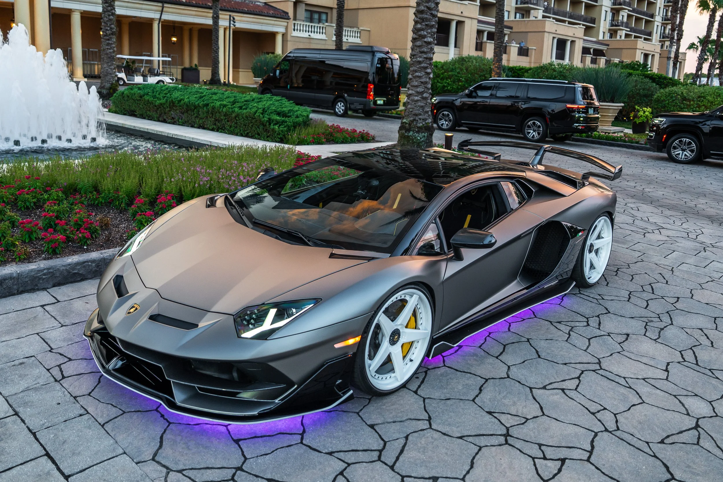 A matte gray Lamborghini sports car parked on a stone-paved driveway in front of a hotel or residential building, with purple underglow lighting beneath the car. The background features landscaped bushes, flowers, palm trees, and several black SUVs.
