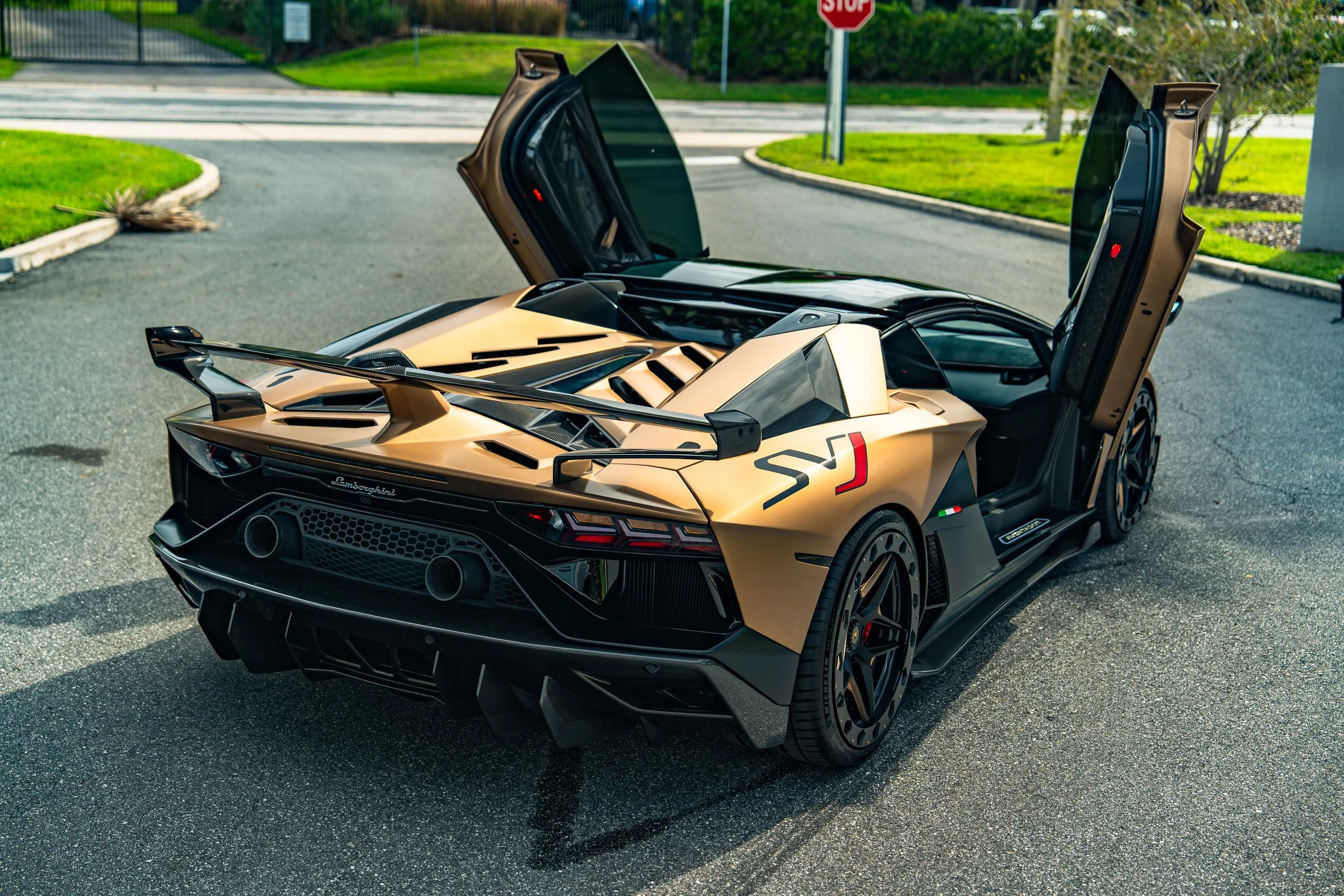 A gold Lamborghini Huracan STO with scissor doors open, showing the interior, parked on a paved road with grass and trees in the background.