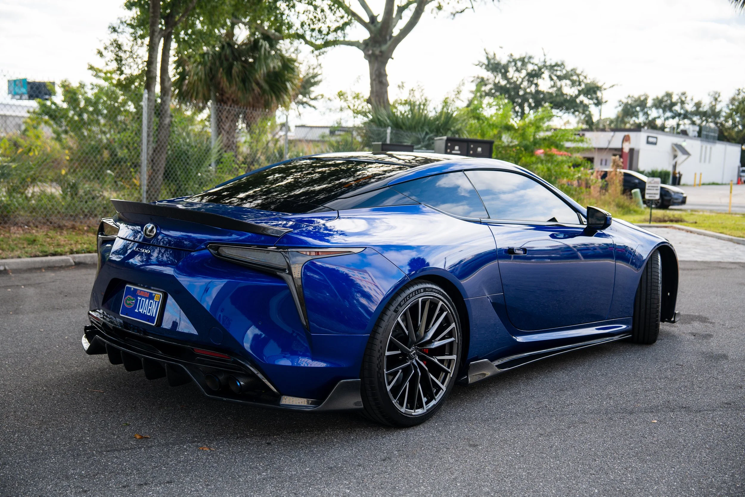 Blue Lexus LC 500 sports car parked on street, rear view, with trees and nearby buildings in the background.