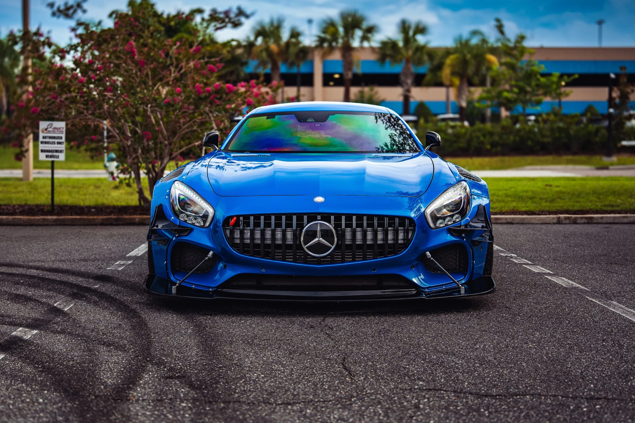 A blue Mercedes-Benz sports car parked in a parking lot with a tree with pink flowers and a building with palm trees in the background.