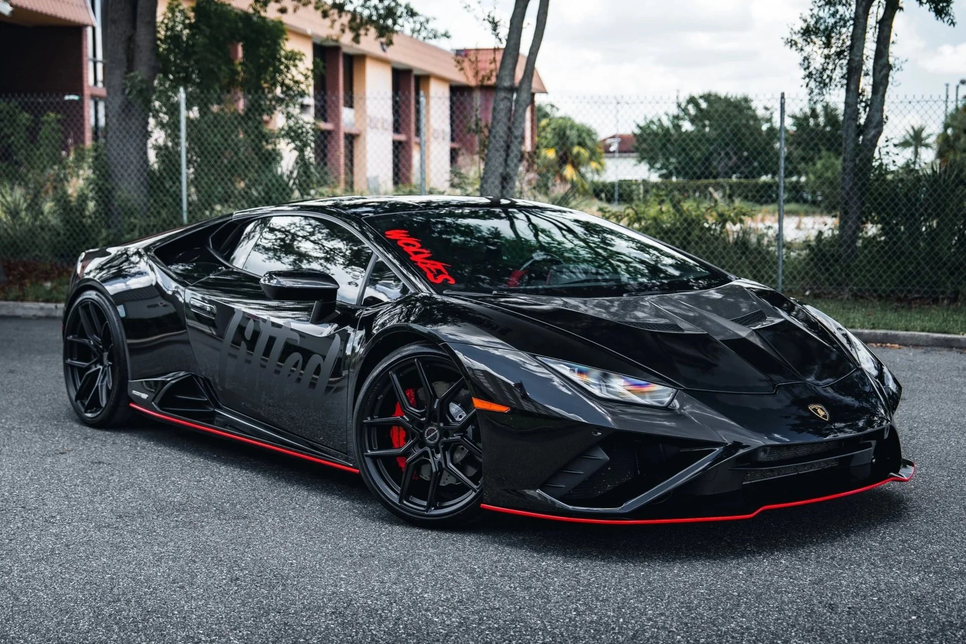 A black Lamborghini sports car parked on a paved road with a chain-link fence and trees in the background.