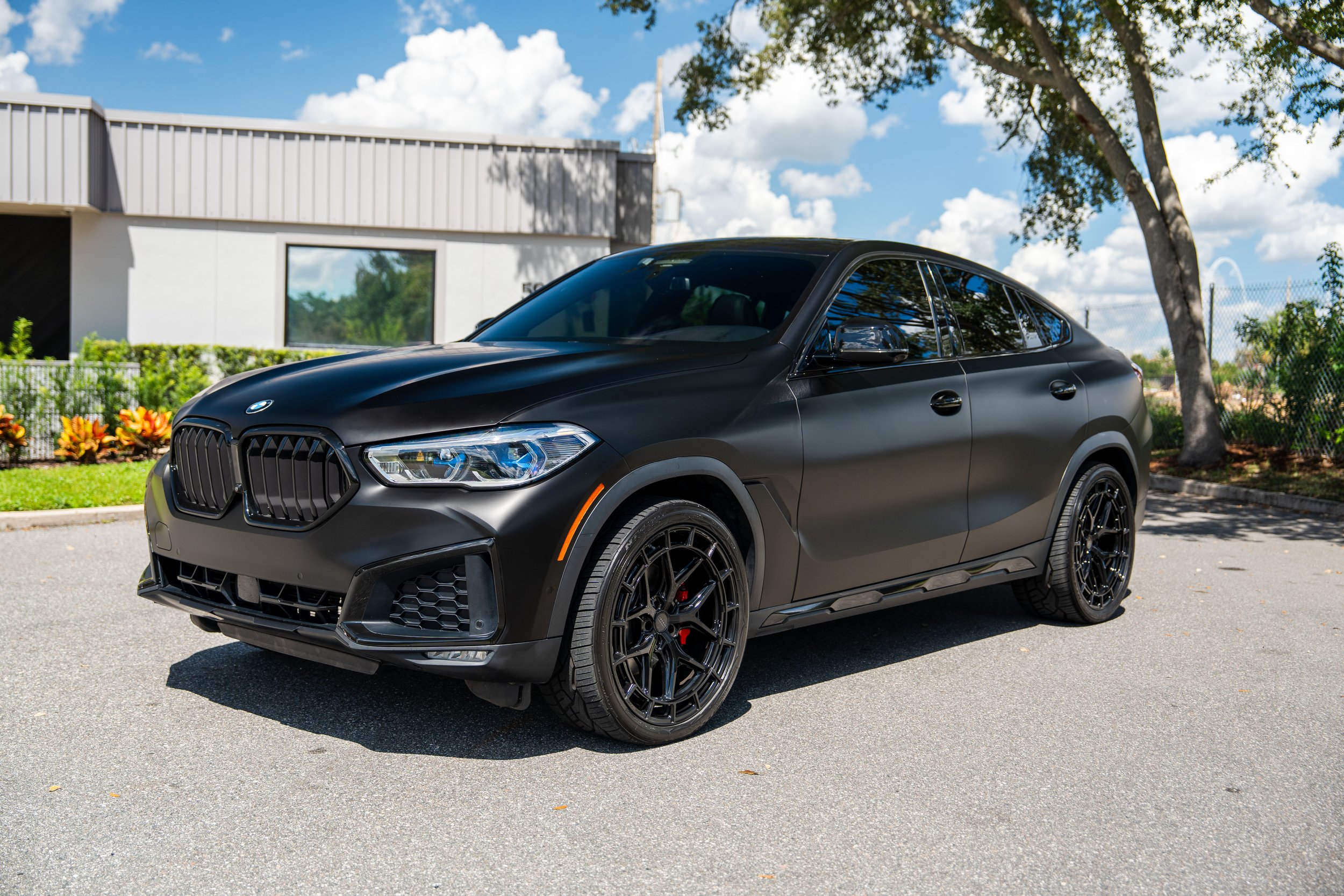 Black BMW SUV parked outdoors on a sunny day with a modern building, trees, and cloudy sky in the background.