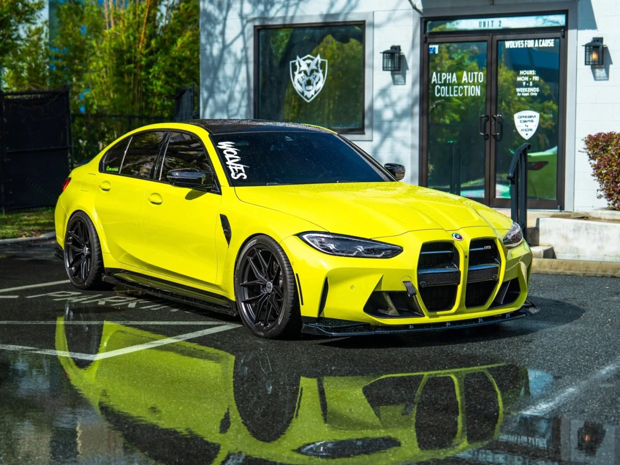 A yellow sports car parked in a lot near a building with a sign that reads "Alpha Auto Collection". The reflection of the car is visible in a wet surface.