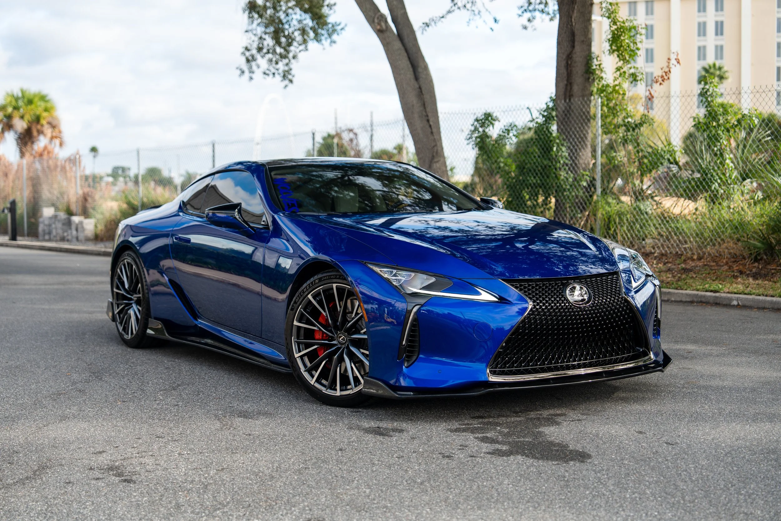Blue Lexus sports car parked on the side of a street with trees and buildings in the background.