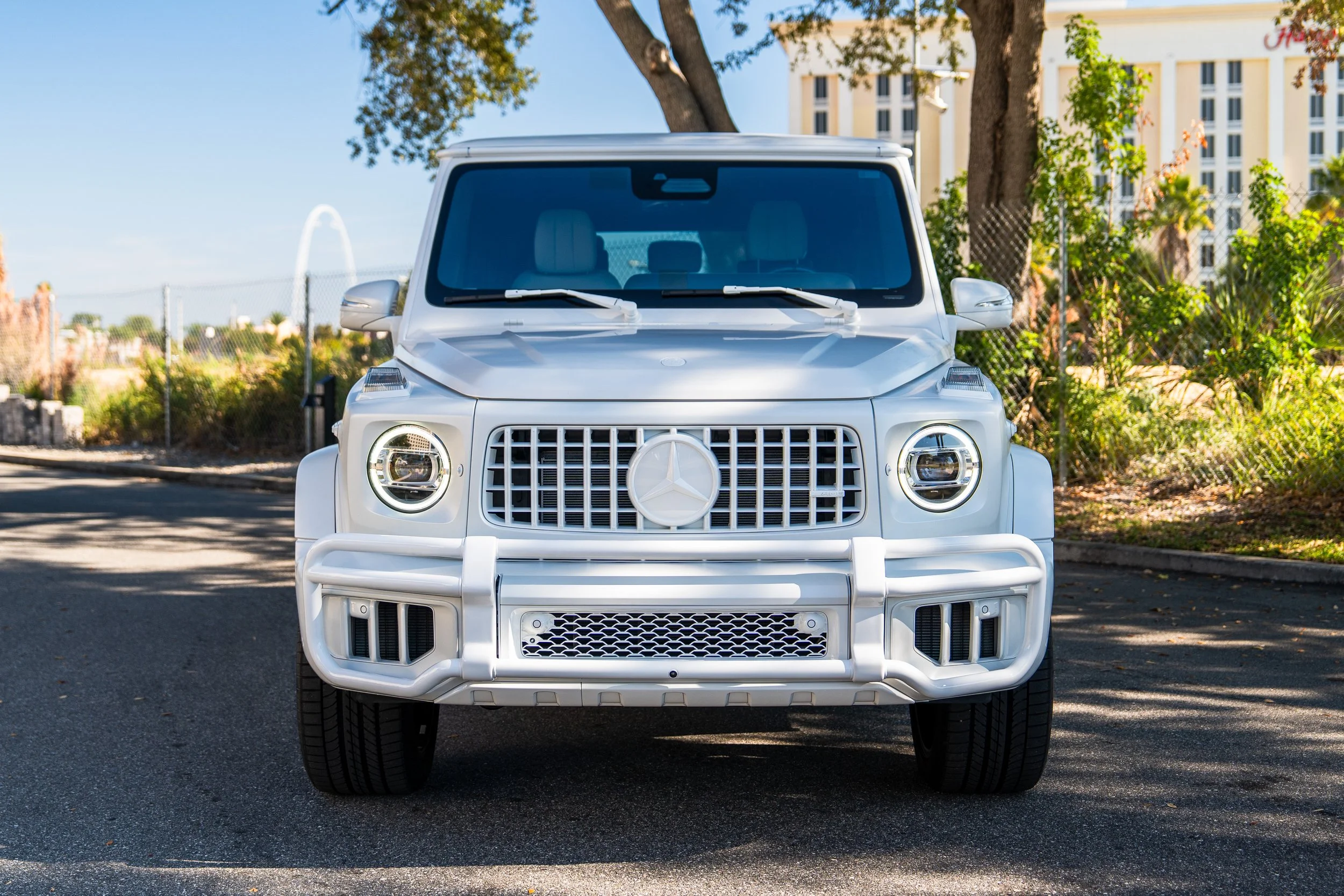 Front view of a white Mercedes-Benz G-class SUV parked on a street, trees, and a fence in the background.