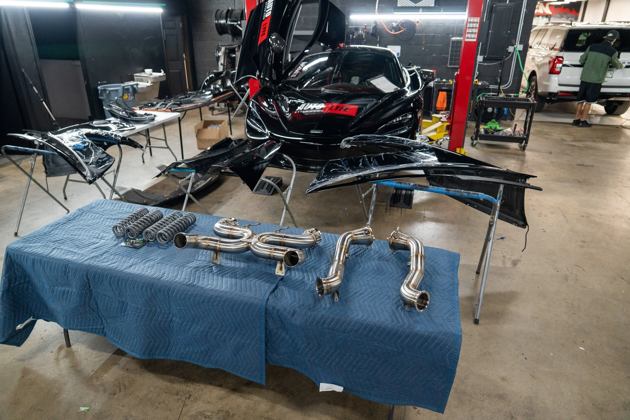 A workshop with a black sports car and various car parts. On a table, there are custom metal exhaust pipes and headers. The scene includes disassembled car body parts, tools, and workbenches, indicating a vehicle modification or repair setting.