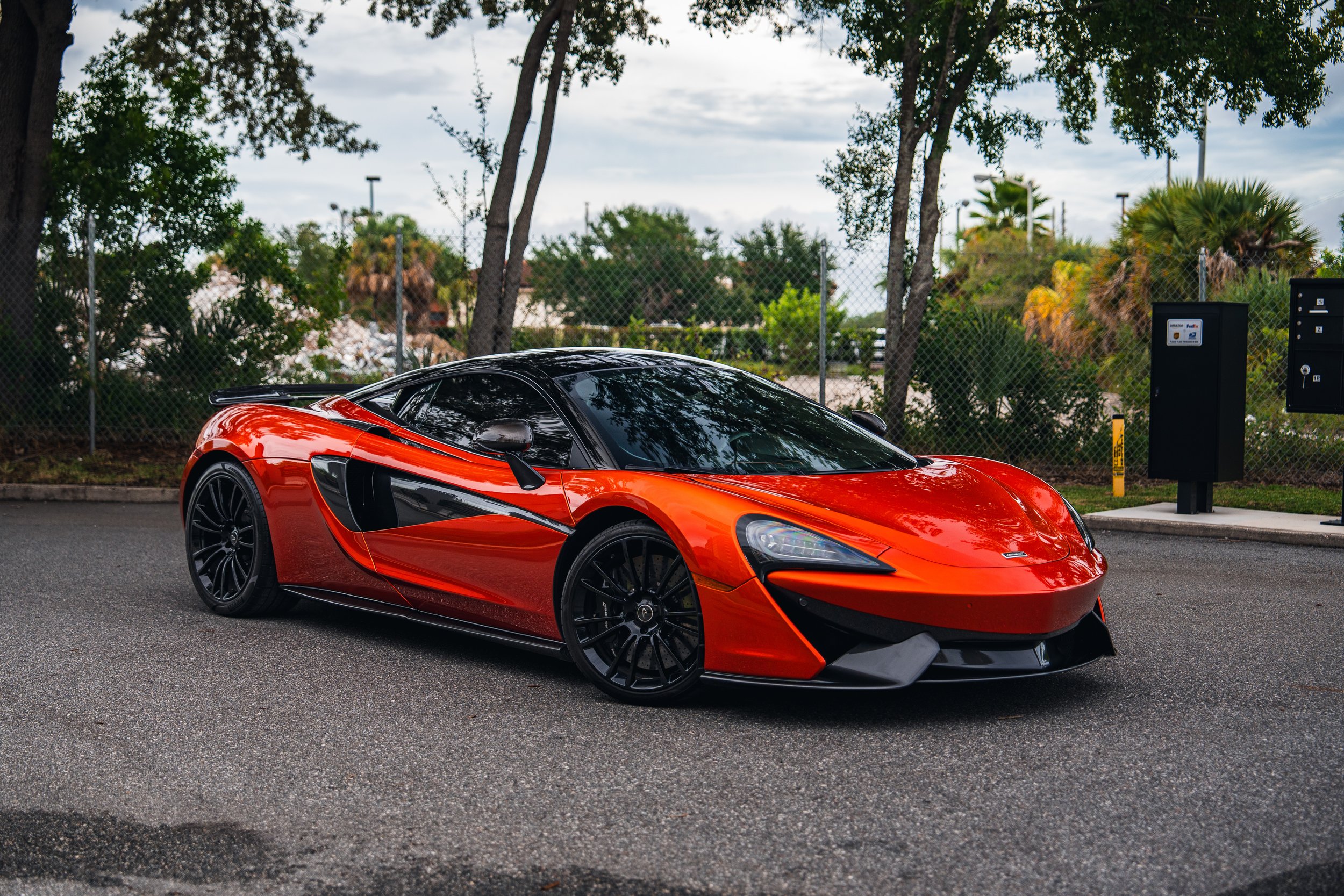 A red and black high-performance sports car parked on a paved surface with trees and a parking lot fence in the background.