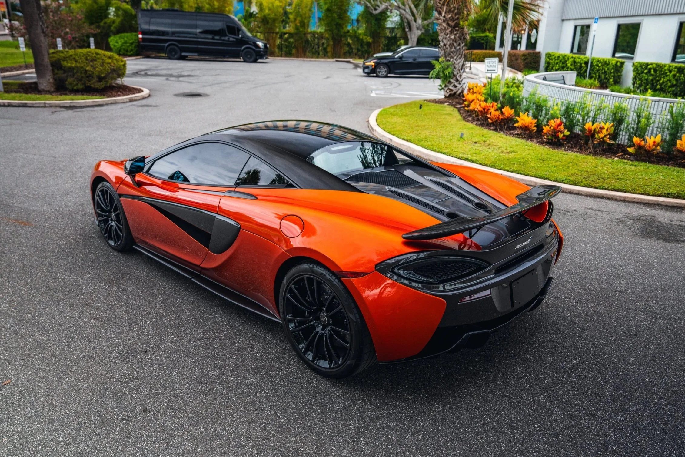A sleek orange sports car with black accents and tinted windows parked in a parking lot near a landscaped area with colorful flowers and palm trees.