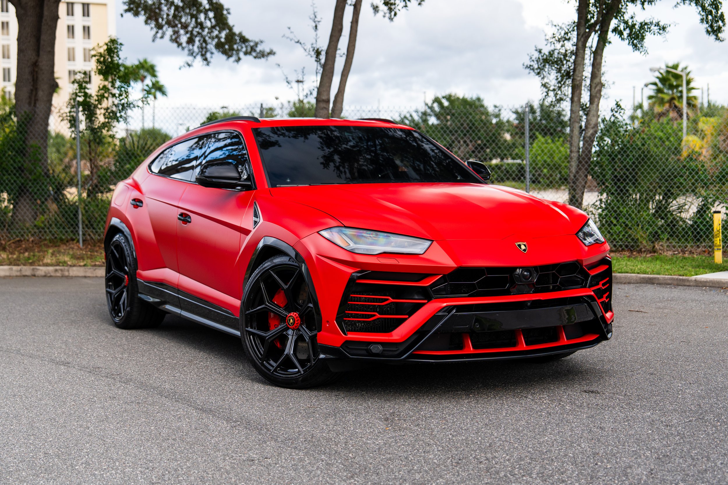 Red Lamborghini Urus parked on a paved surface with a chain-link fence and palm trees in the background.