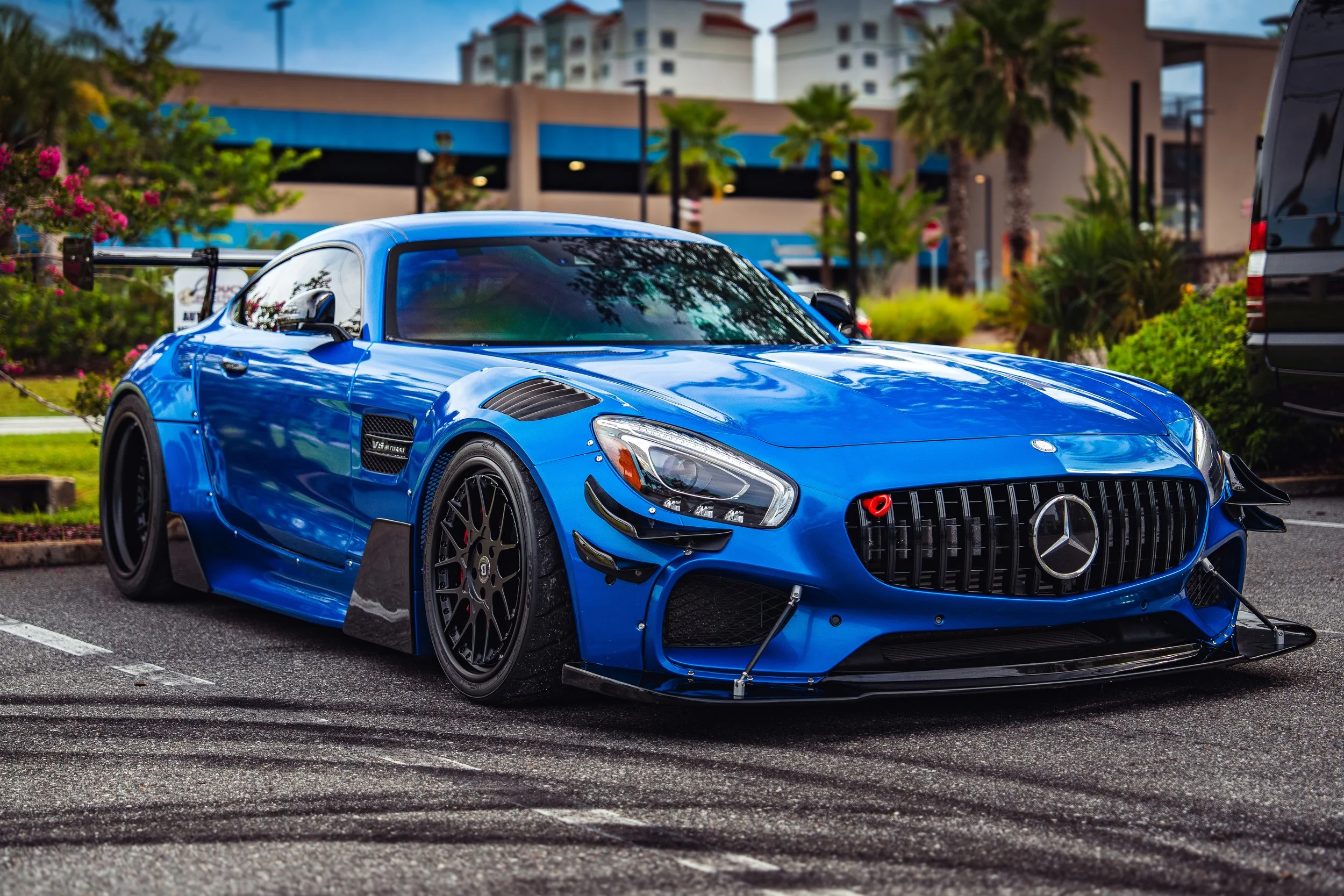 A blue Mercedes-Benz sports car with racing modifications parked in a parking lot with buildings and trees in the background.