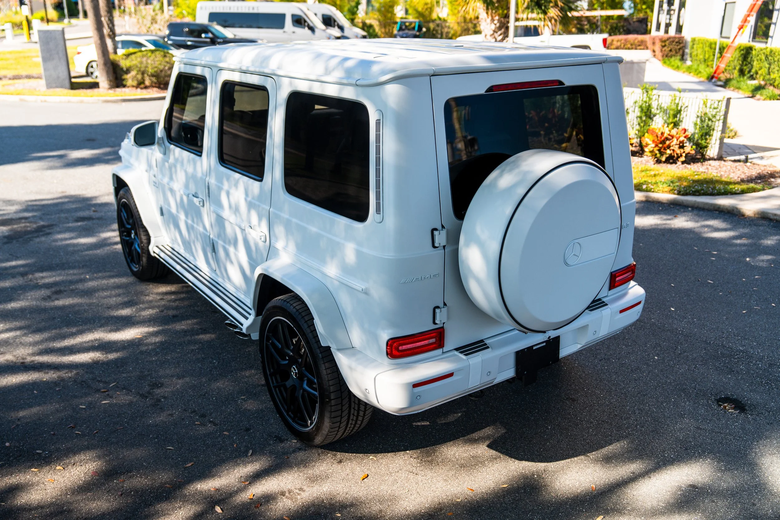 White Mercedes-Benz G-Class SUV parked on a street, with trees casting shadows on the vehicle and a building with plants in the background.