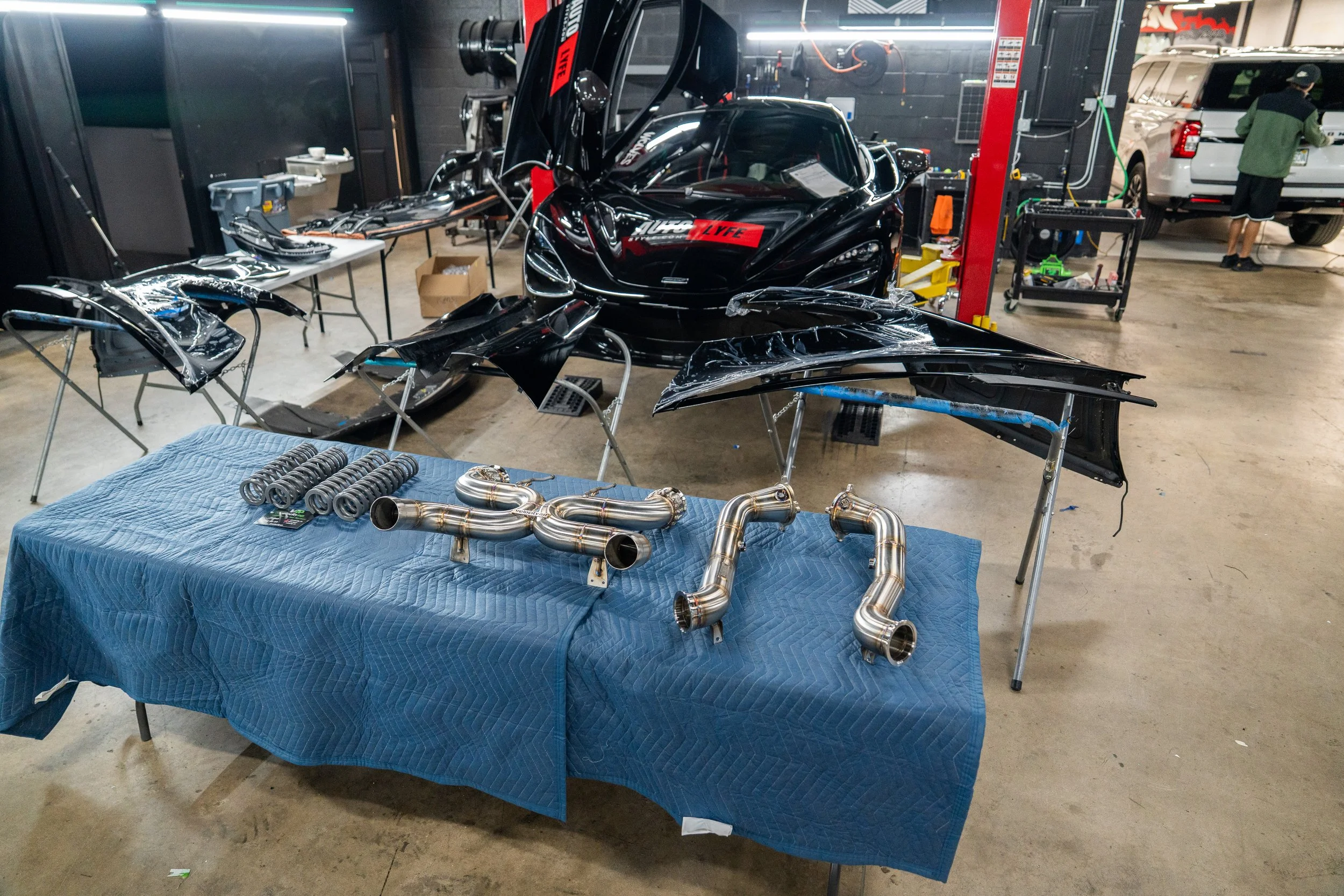 A workshop with car parts including black body panels and an exhaust system laid out on tables. A sleek black sports car is positioned behind the tables, and another vehicle is visible in the background.