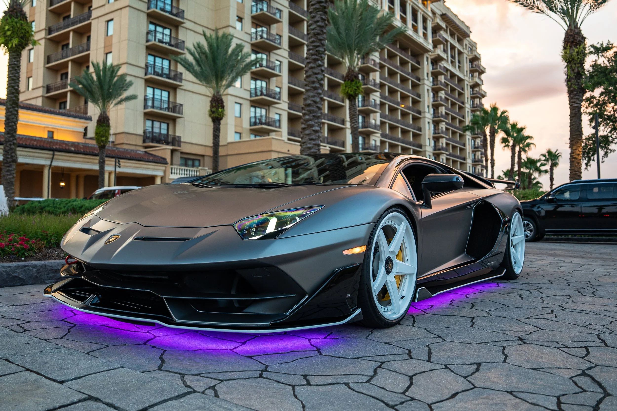 A matte black Lamborghini sports car with neon purple underglow lights parked on a cobblestone driveway, with palm trees and a multi-story apartment building in the background during sunset.