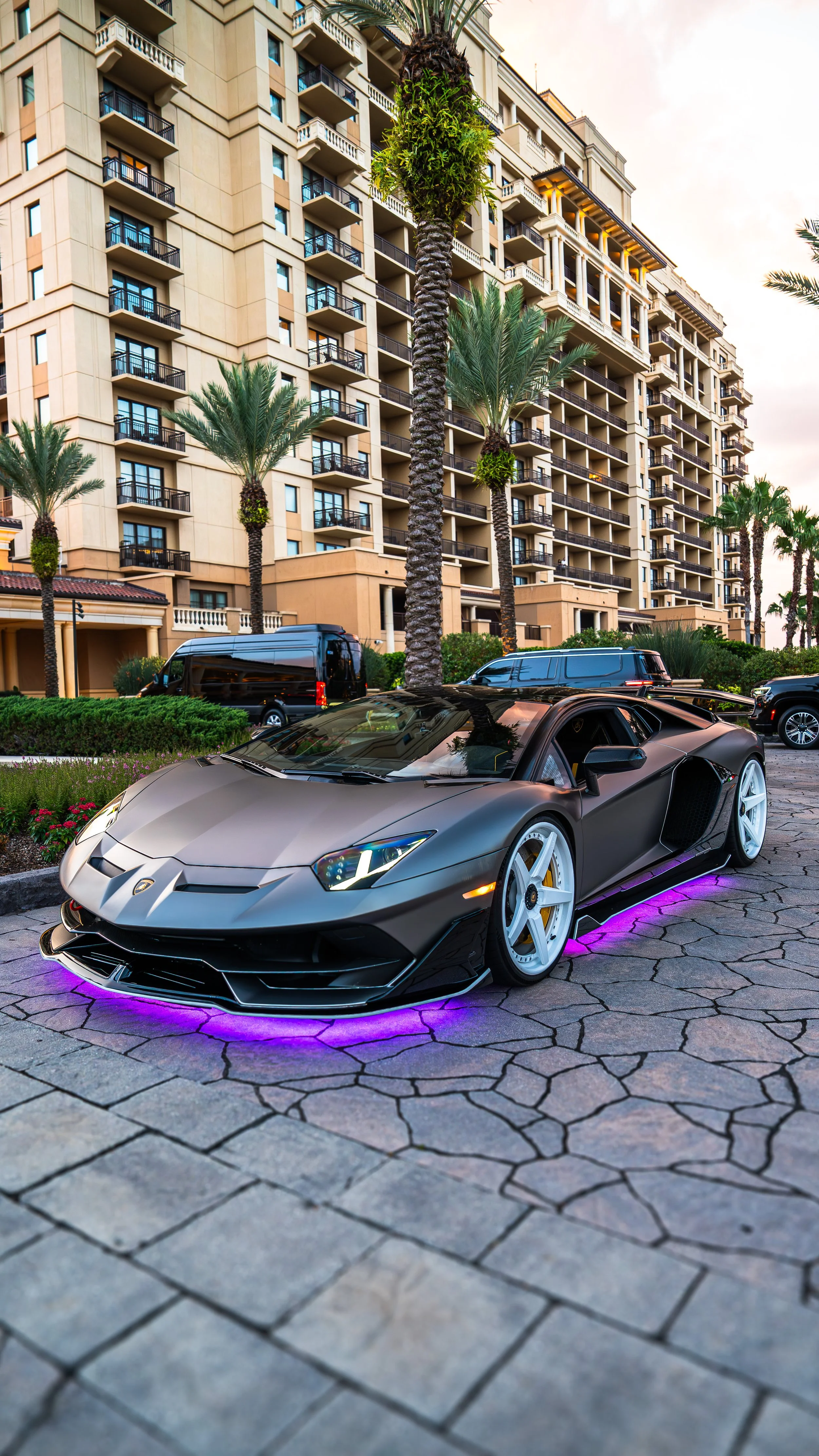 A matte black Lamborghini supercar with purple underglow lights parked in a lot outside a multi-story beige apartment building with balconies. Tall palm trees line the area, and a black van and SUV are parked nearby during sunset.