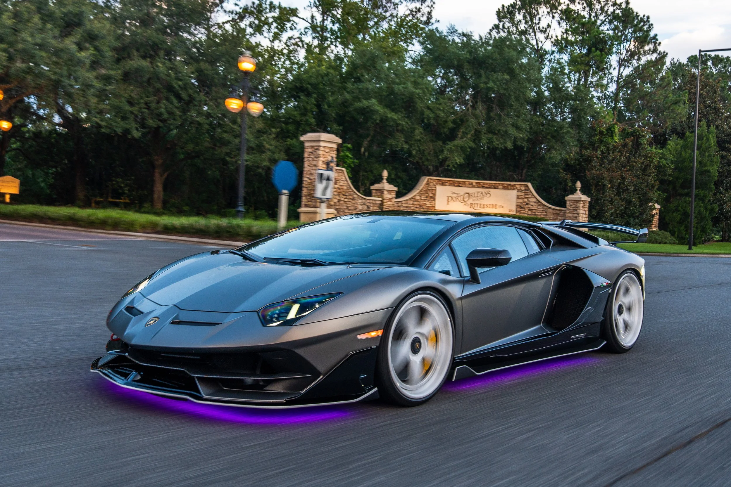 A black Lamborghini supercar driving on a road with purple underglow lights, trees, and a sign that reads 'The Orleans Riverside' in the background.