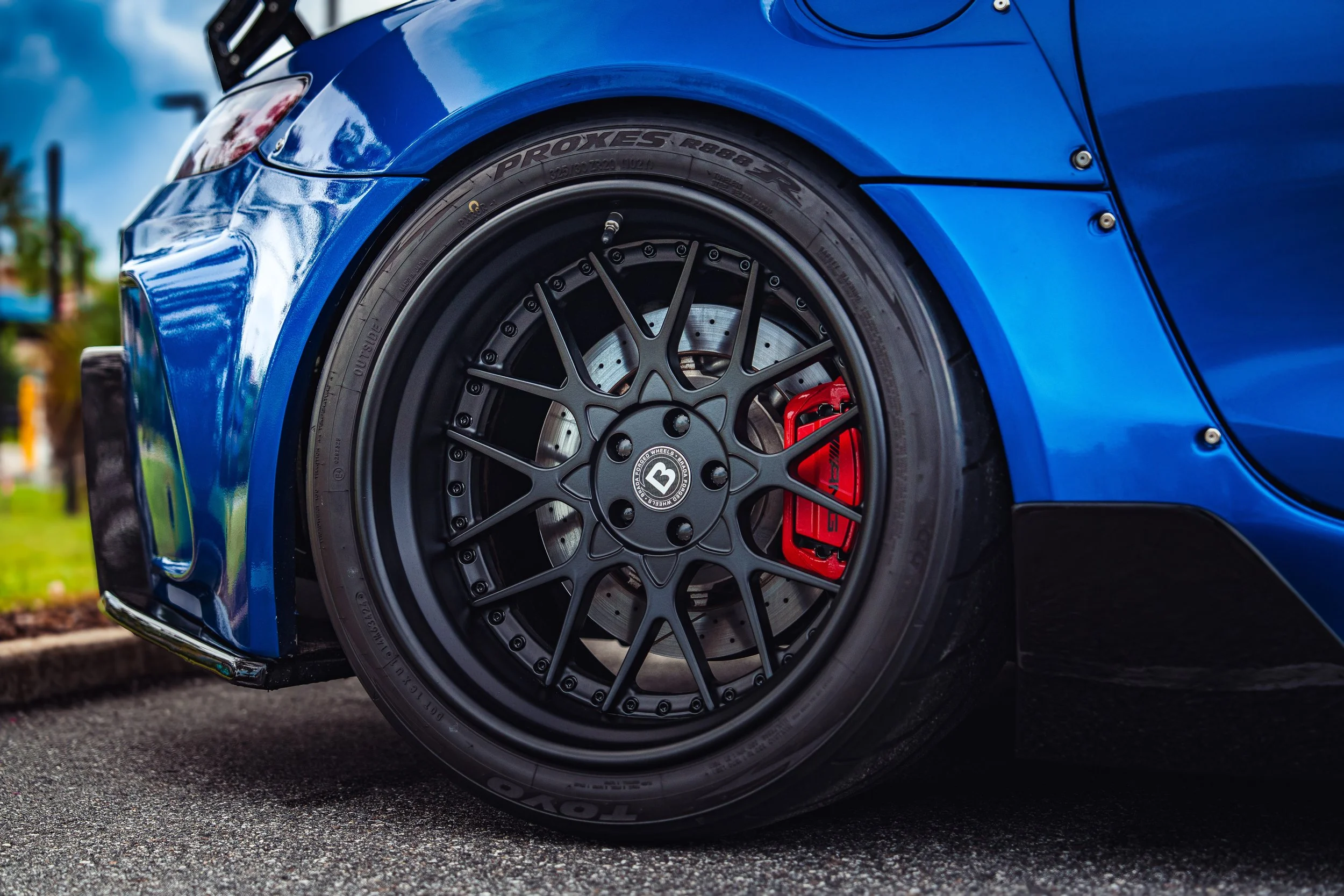 Close-up of a blue sports car's front wheel featuring a black rim, red brake caliper, and drilled brake disc.