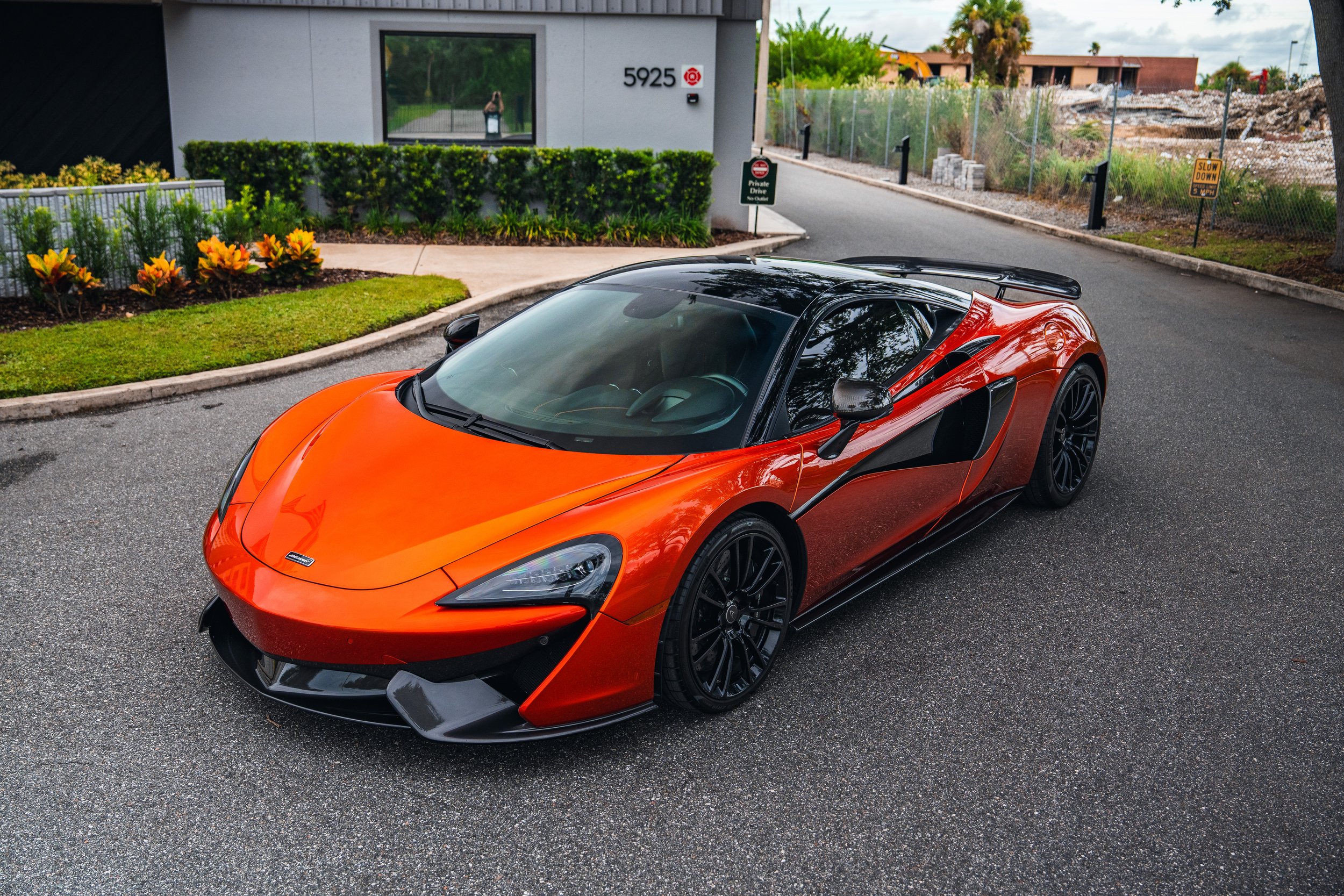 Orange McLaren sports car parked on the street near a modern building with greenery and flowers.