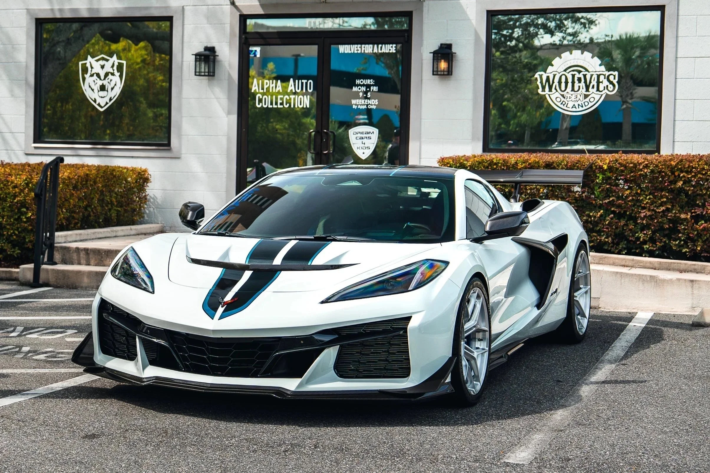 A white sports car with black and blue racing stripes parked outside a car dealership with signage that reads 'Alpha Auto Collection' and 'Wolves Orlando', with a logo of a wolf's head.
